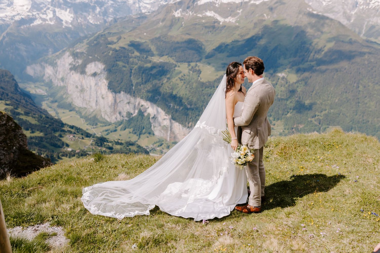 A couple embraces on a scenic mountain meadow with dramatic Swiss Alps peaks in the background on their wedding day.