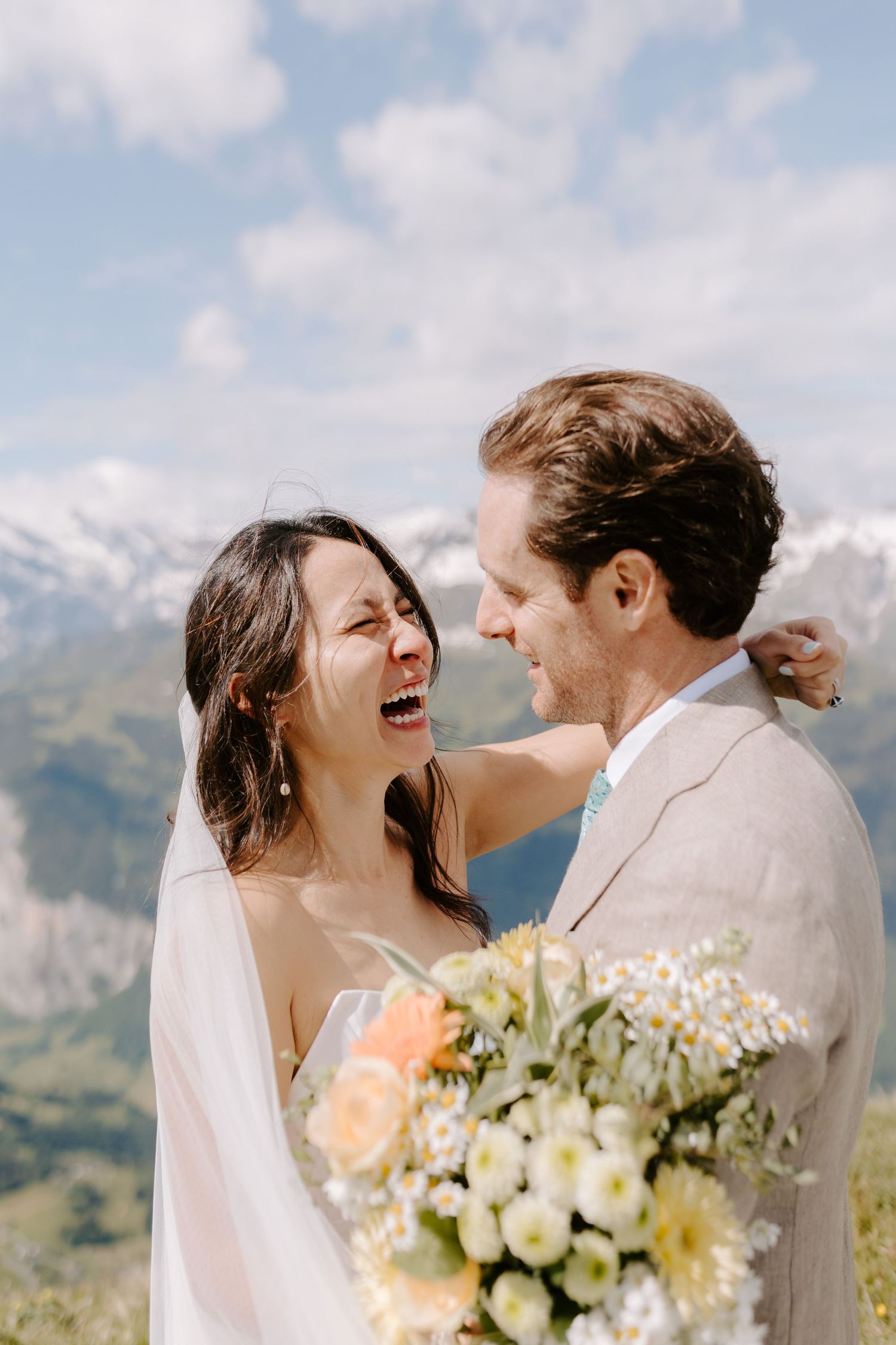 Happy newlywed couple sharing a joyful moment against a mountainous backdrop with a bridal bouquet of mixed flowers.