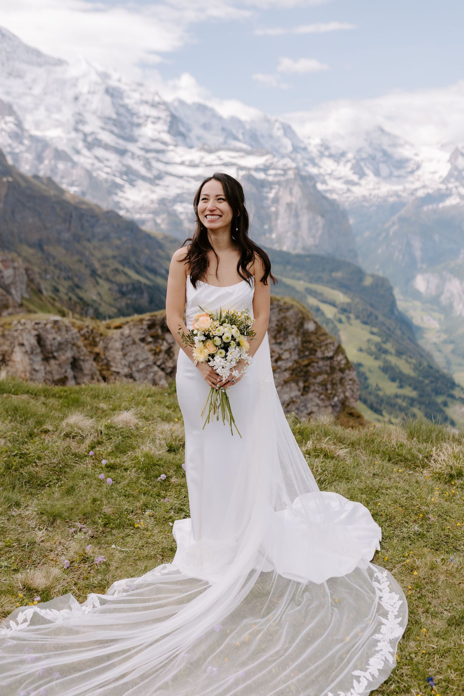 A white wedding dress flows elegantly against a mountainous alpine backdrop with snow-capped peaks.