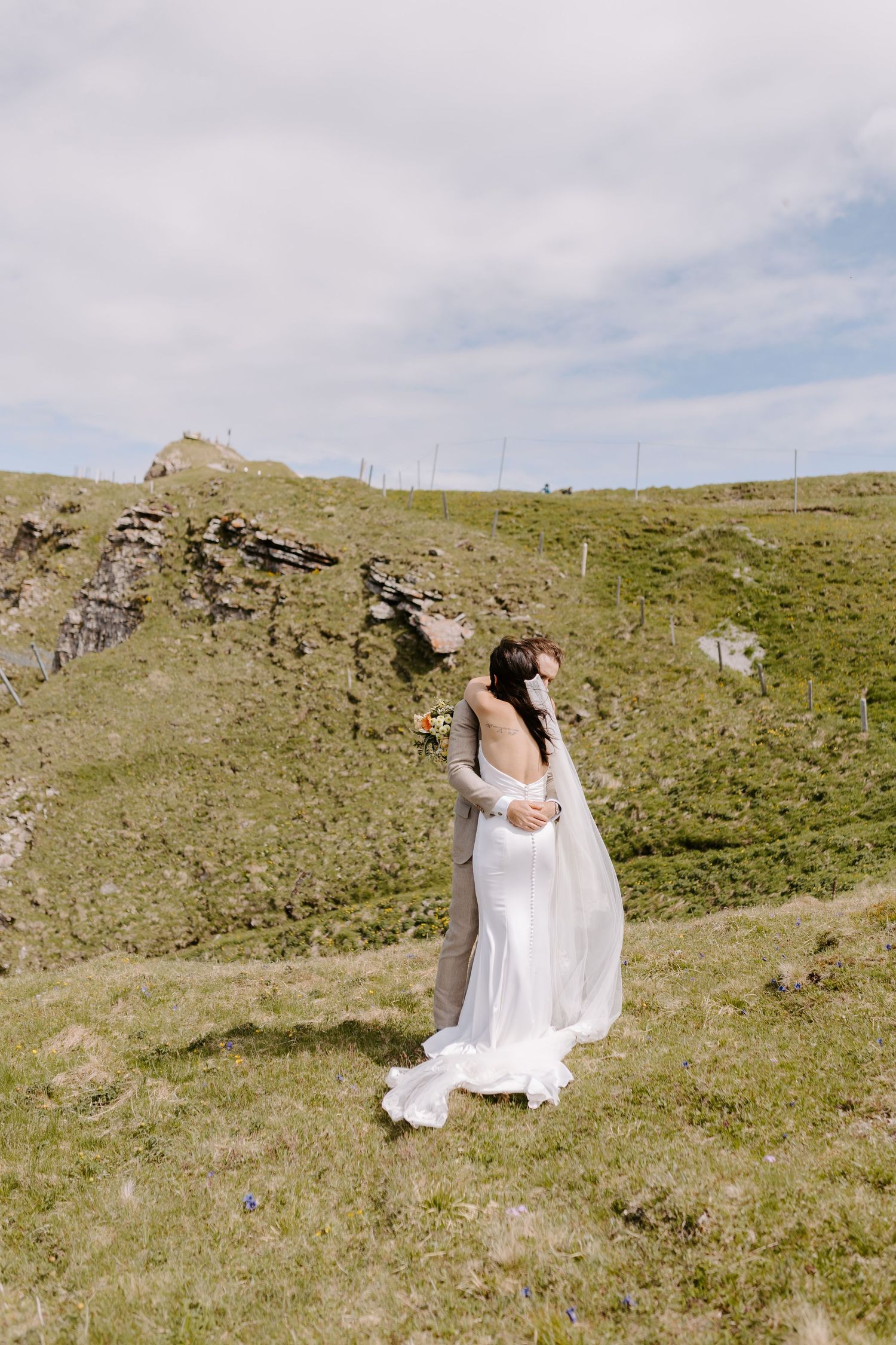 A bride in a flowing white wedding dress stands on a grassy hillside against dramatic rocky cliffs and cloudy sky.
