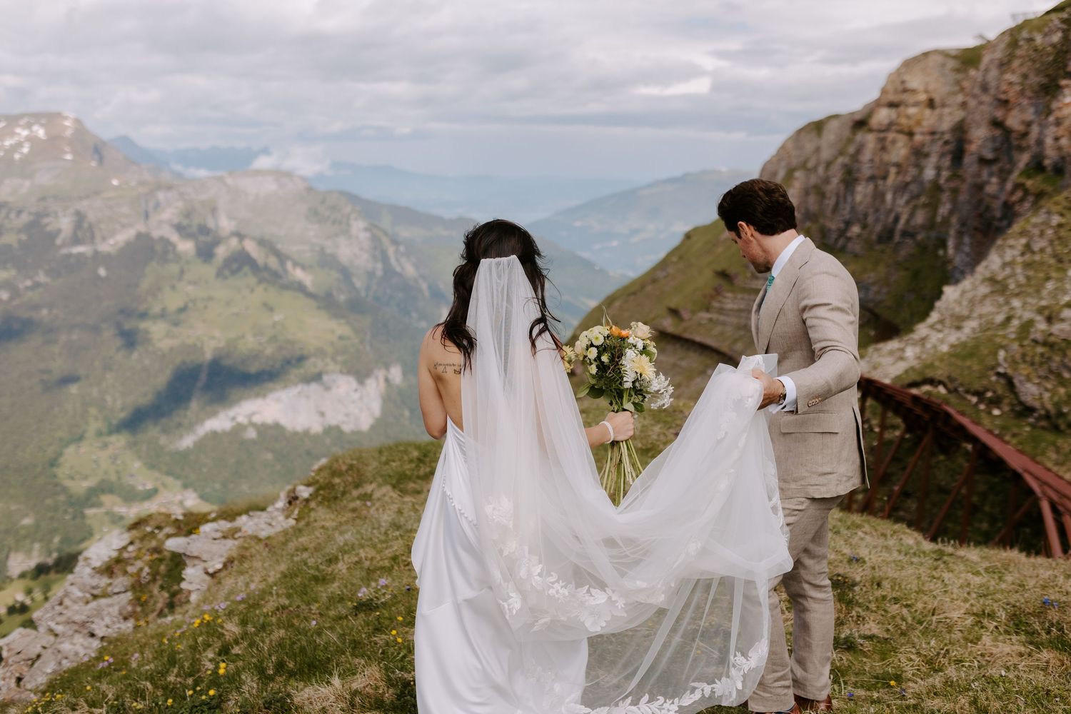 A couple in wedding attire stands on a scenic mountain peak with dramatic Dolomites landscape in the background.