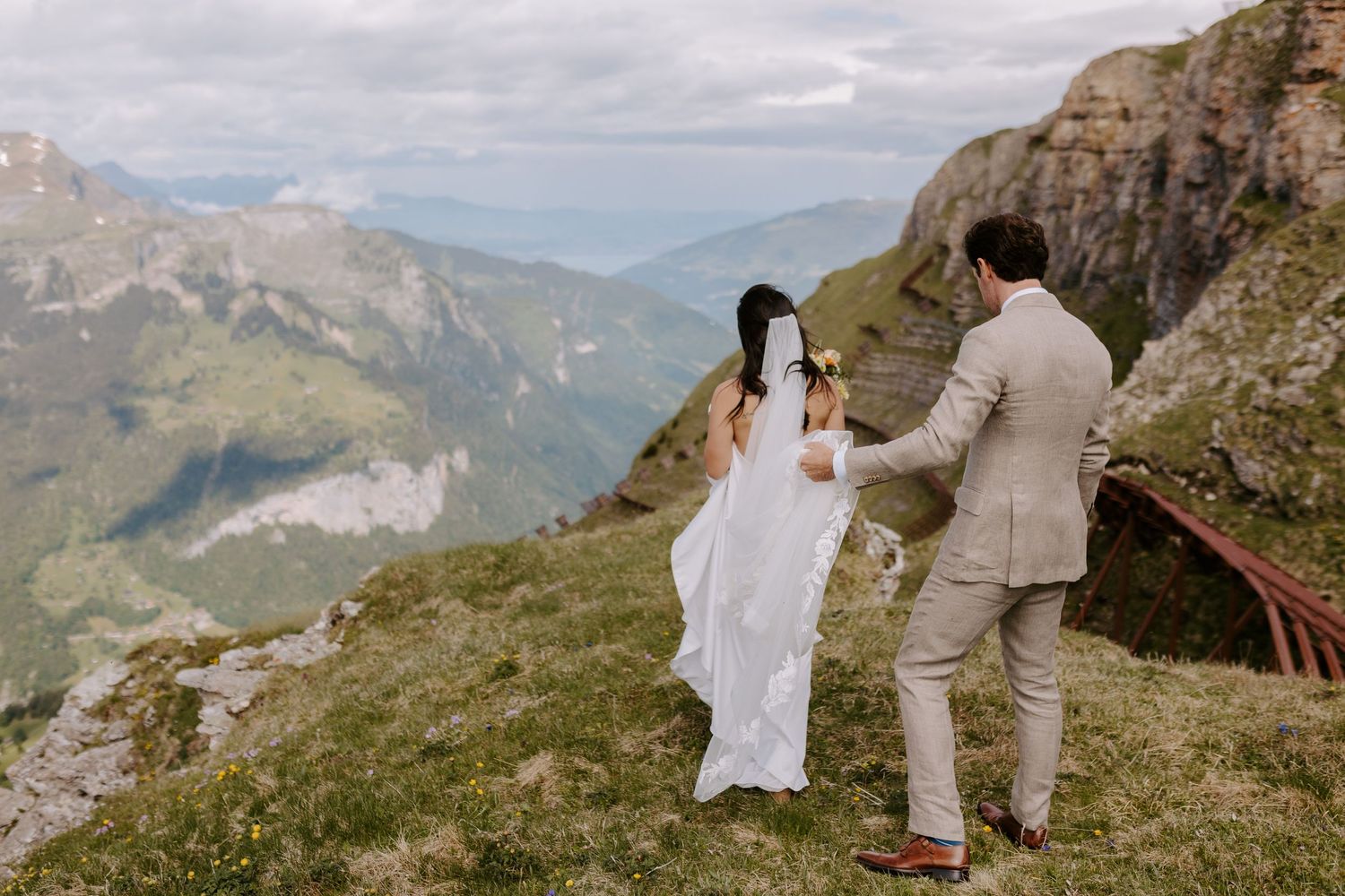 A couple in wedding attire walks hand in hand along a scenic mountain ridge overlooking a dramatic alpine landscape.