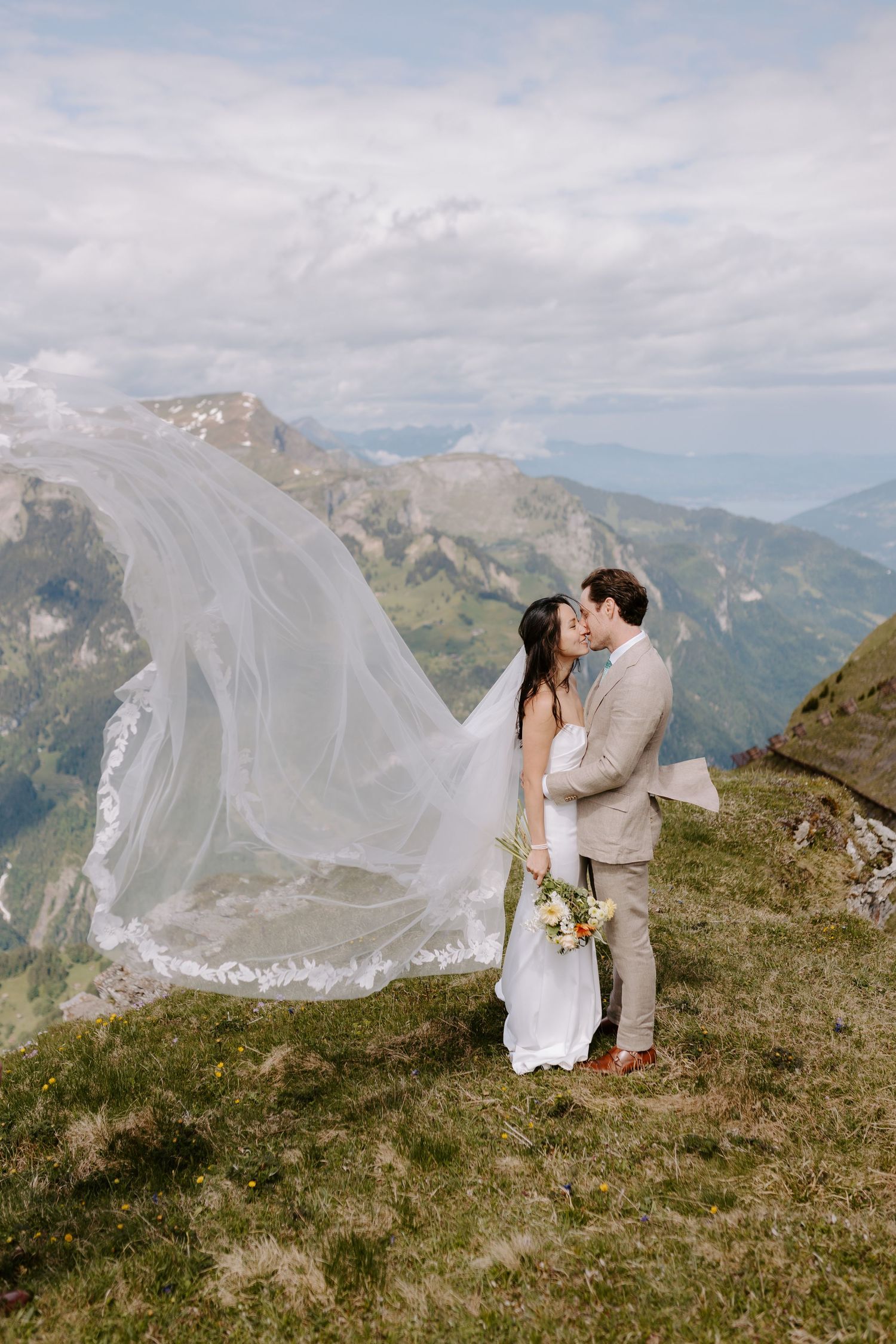 A couple in wedding attire share a romantic moment on a mountaintop as a long veil flows dramatically in the wind.