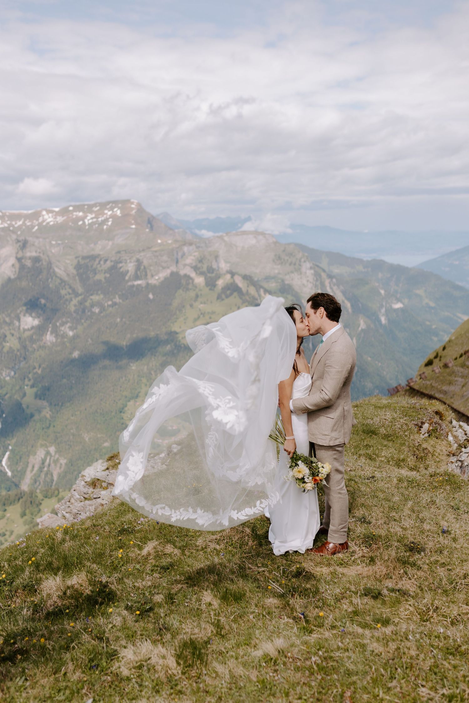 A couple shares an intimate moment on a mountaintop overlooking majestic Alpine peaks and valleys in the background.