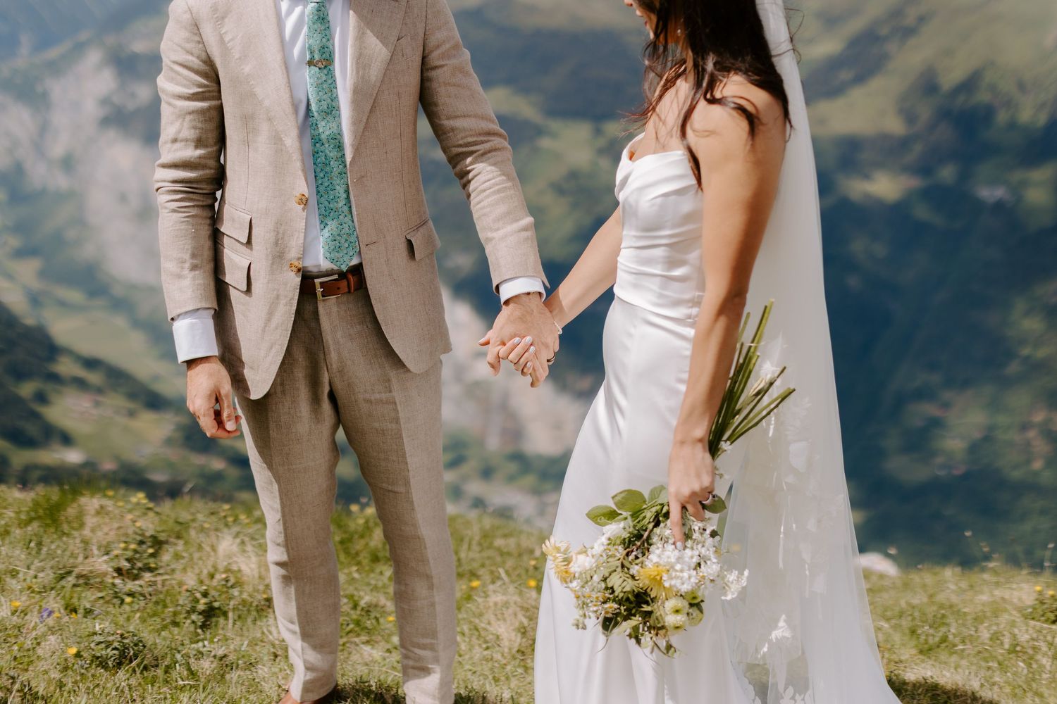 Newlywed couple holding hands on mountain meadow with scenic alpine peaks in background during intimate wedding ceremony.
