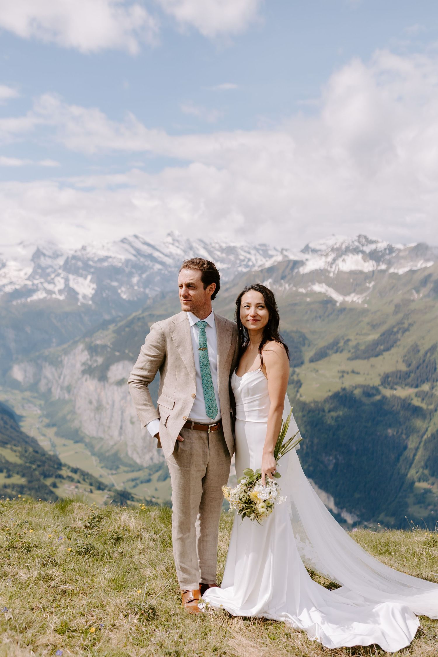 A wedding couple poses against a backdrop of snow-capped mountains in the Alps with blue skies and clouds.