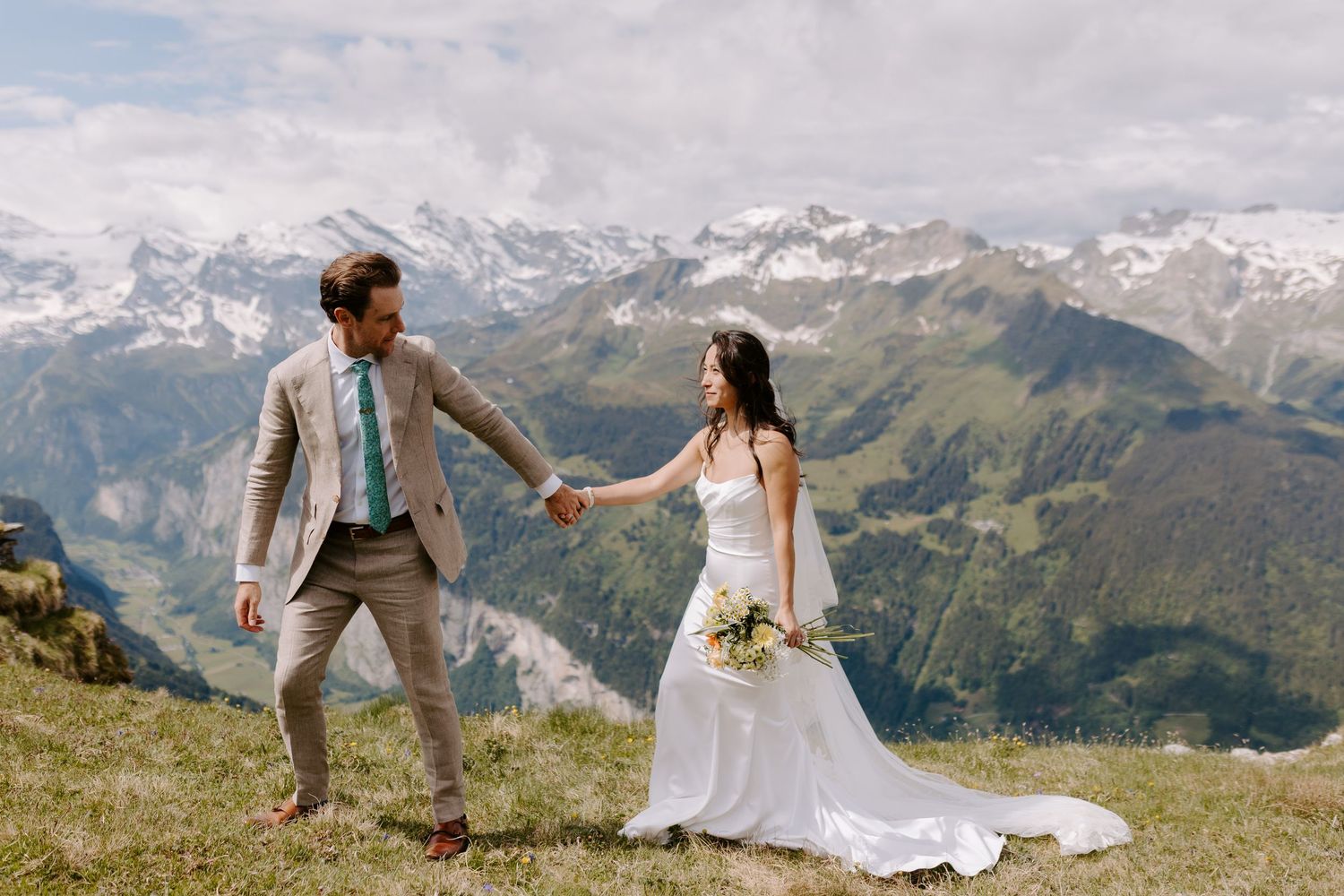 A couple holds hands on a grassy mountain summit with snow-capped Alpine peaks in the background.