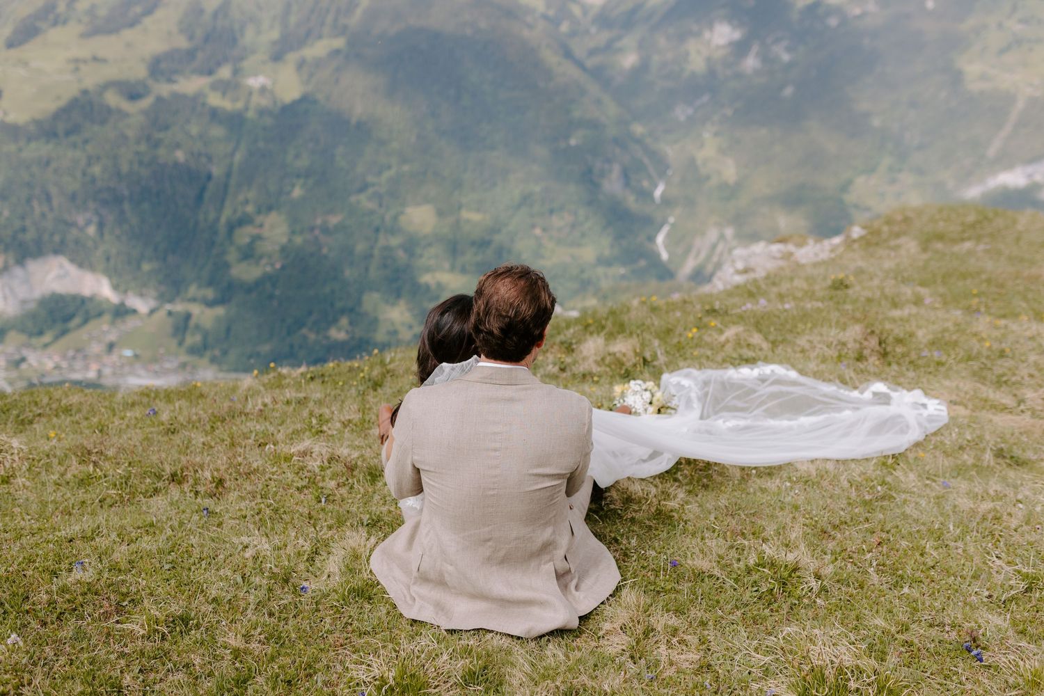 Someone sits on a grassy mountain slope with a flowing white fabric beside them, overlooking a scenic mountain landscape.