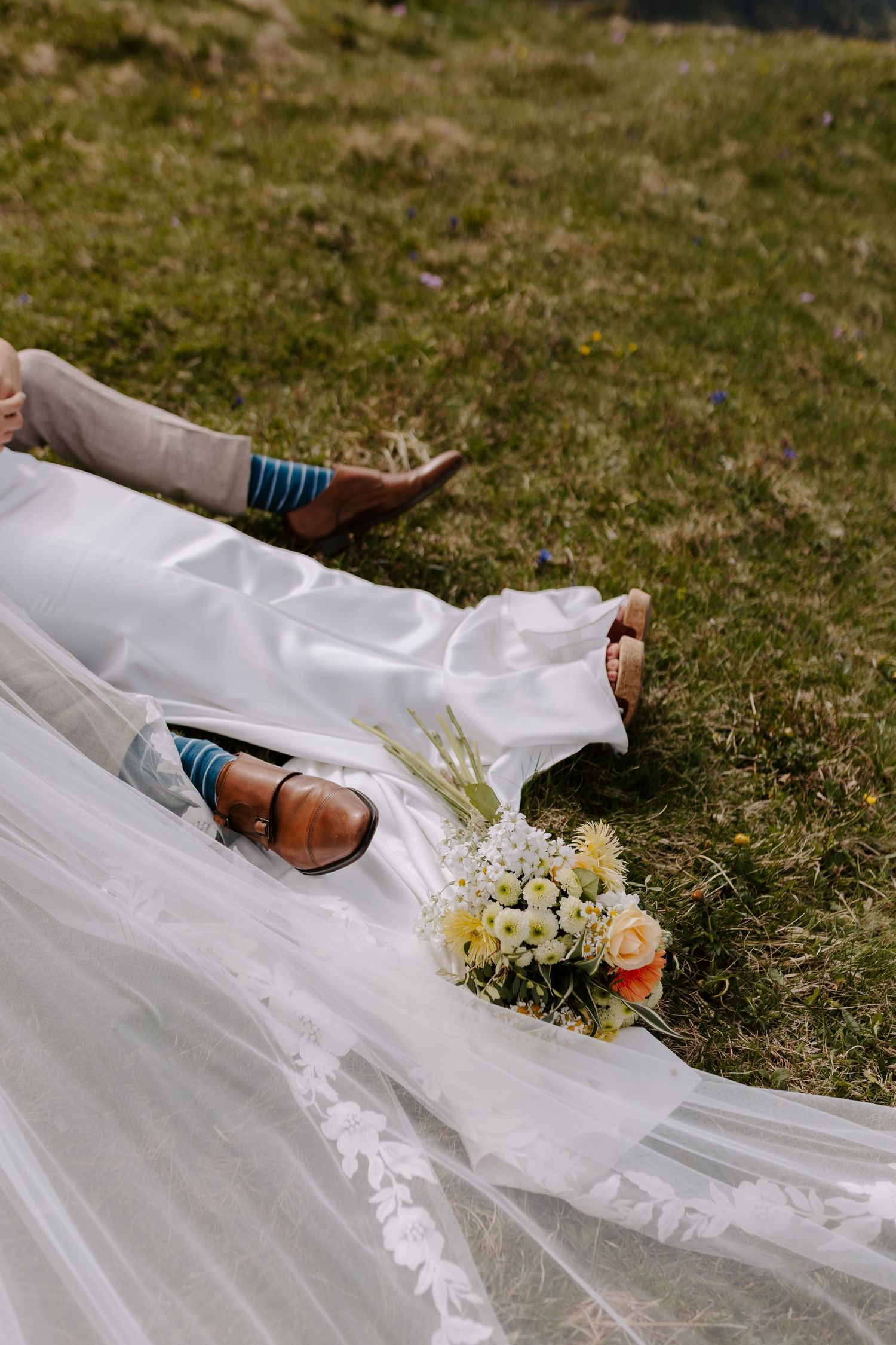 White wedding dress and bridal shoes laid on grass with delicate bouquet of yellow and white flowers.