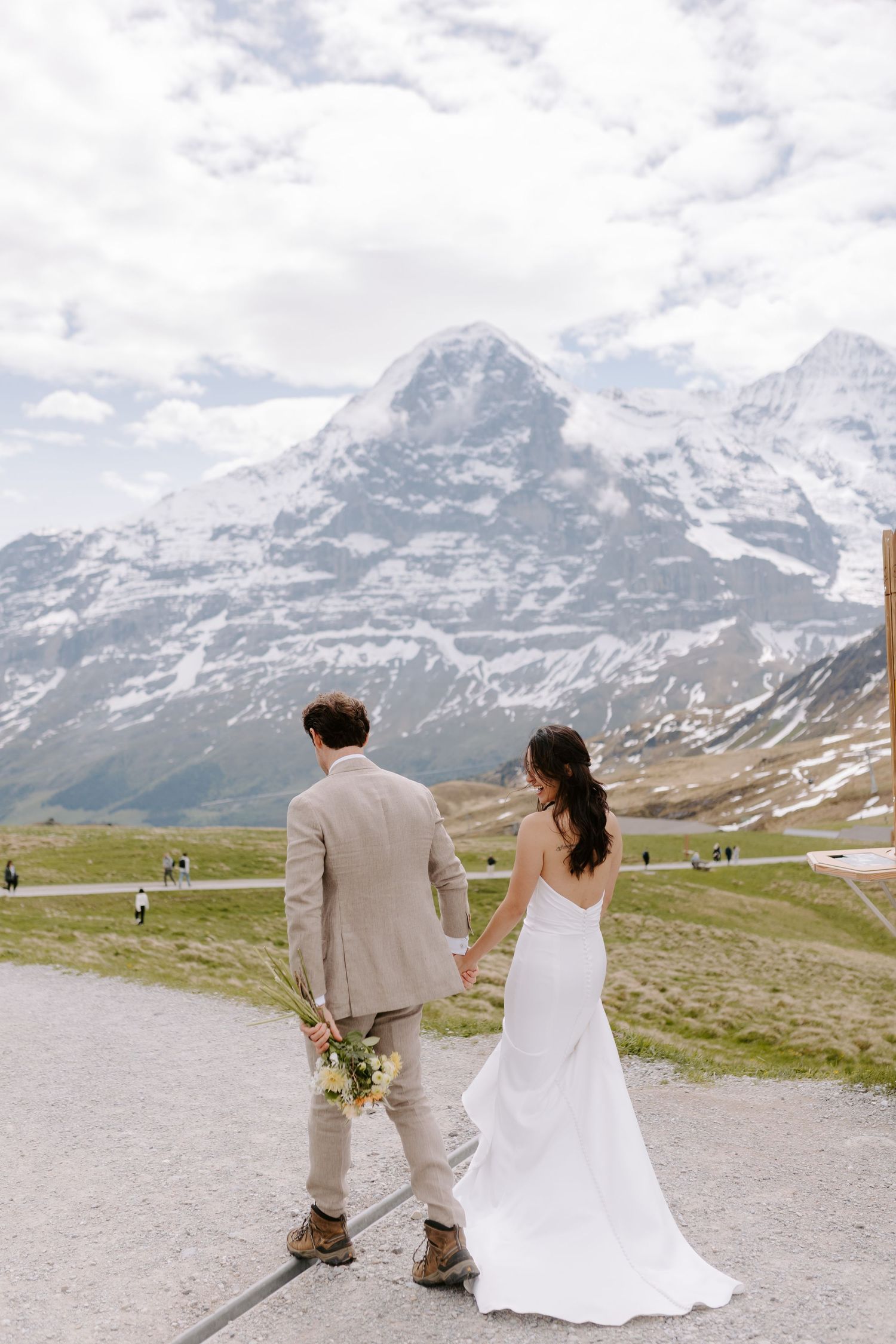 A couple in wedding attire walks along a mountain path with the Swiss Alps and Eiger peak in the background.