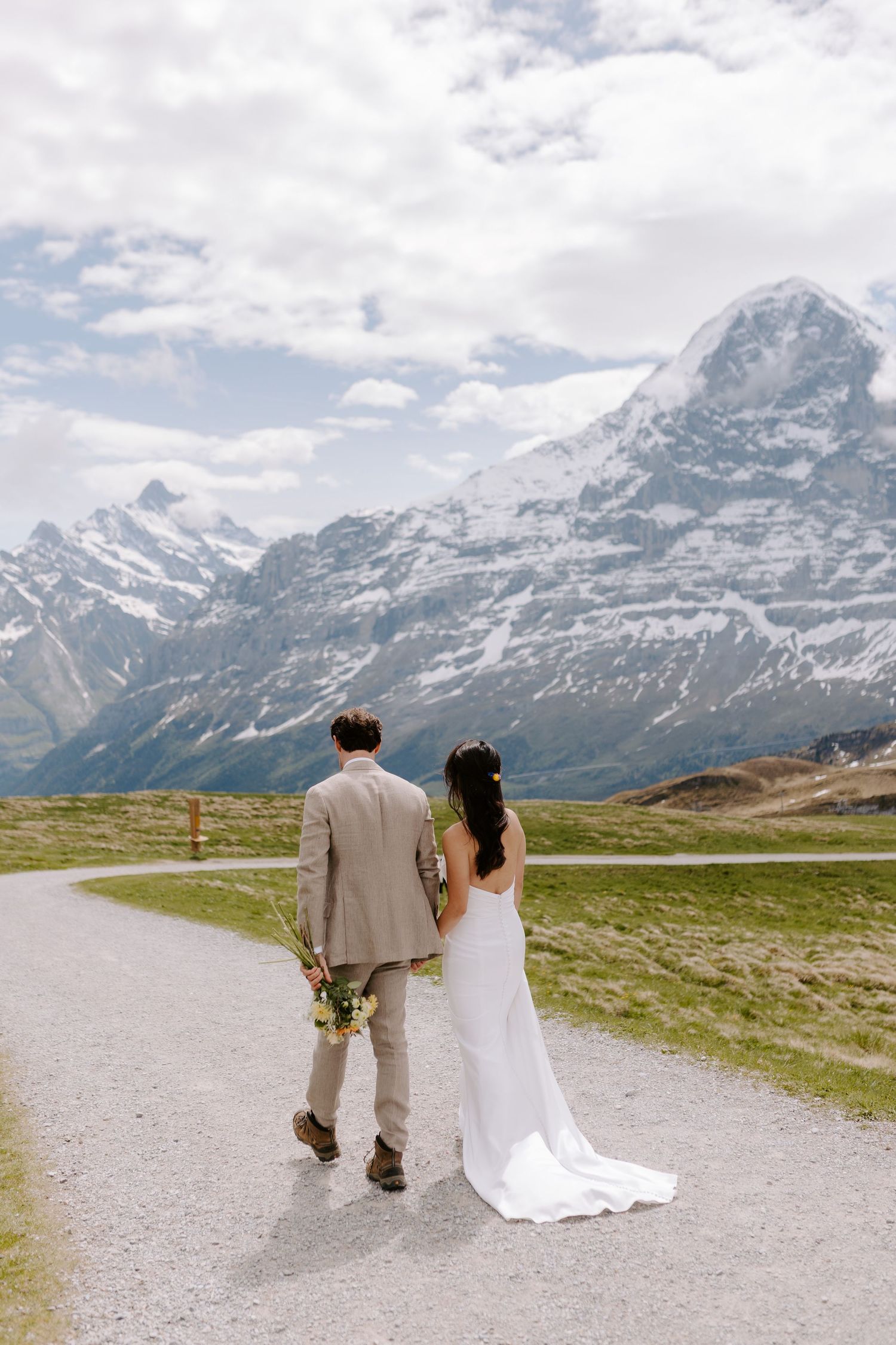 A couple walking along a mountain path with snow-capped Alps peaks towering in the background on their wedding day.