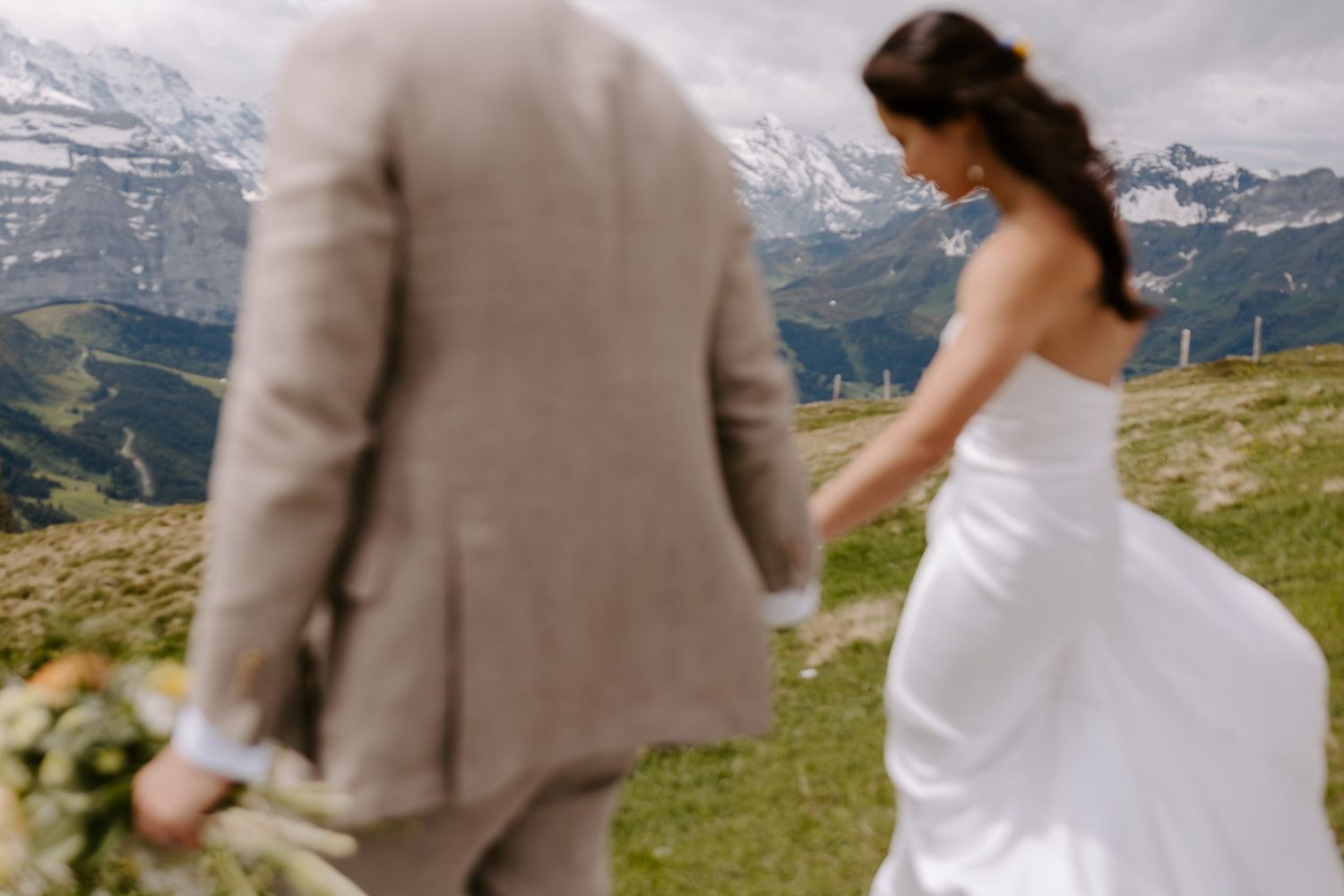 A couple holds hands on a grassy mountainside with snow-capped peaks in the distance during a wedding ceremony.