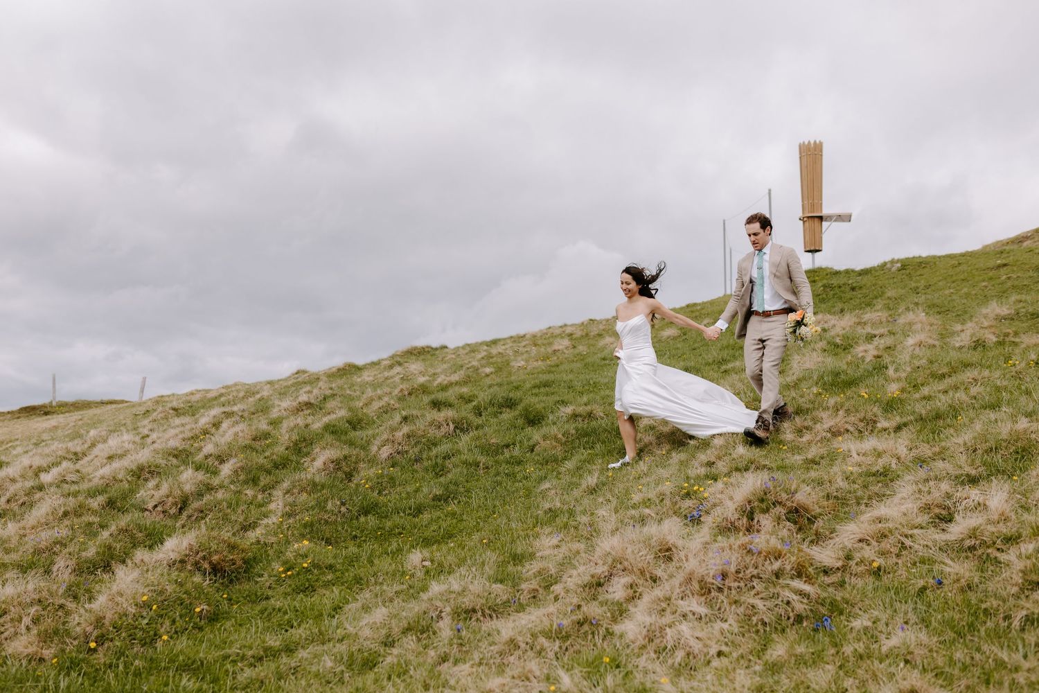 A couple on their wedding day runs joyfully together across a green hillside under an overcast sky.