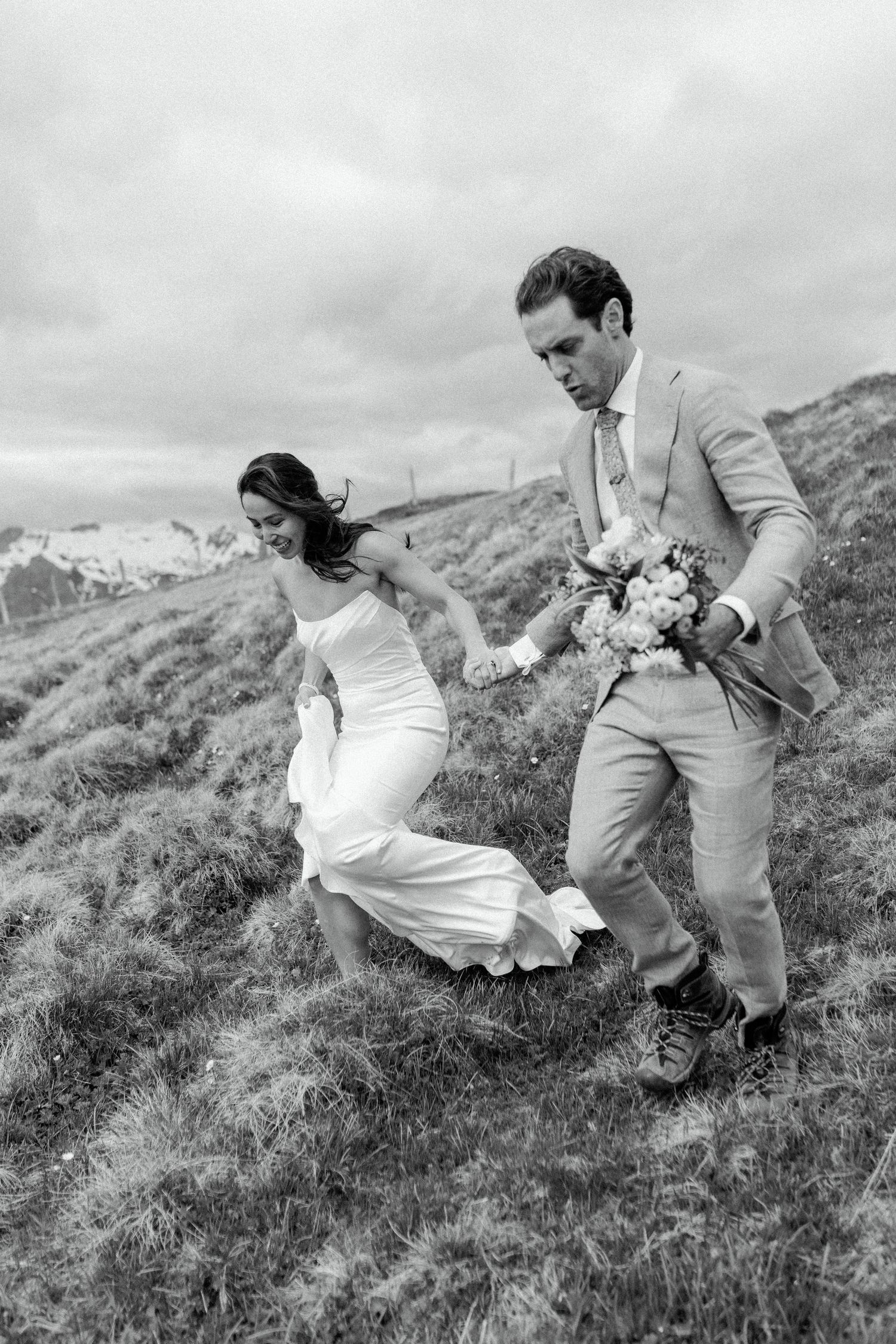 A couple enjoys a playful moment running on a grassy hillside in black and white wedding attire.