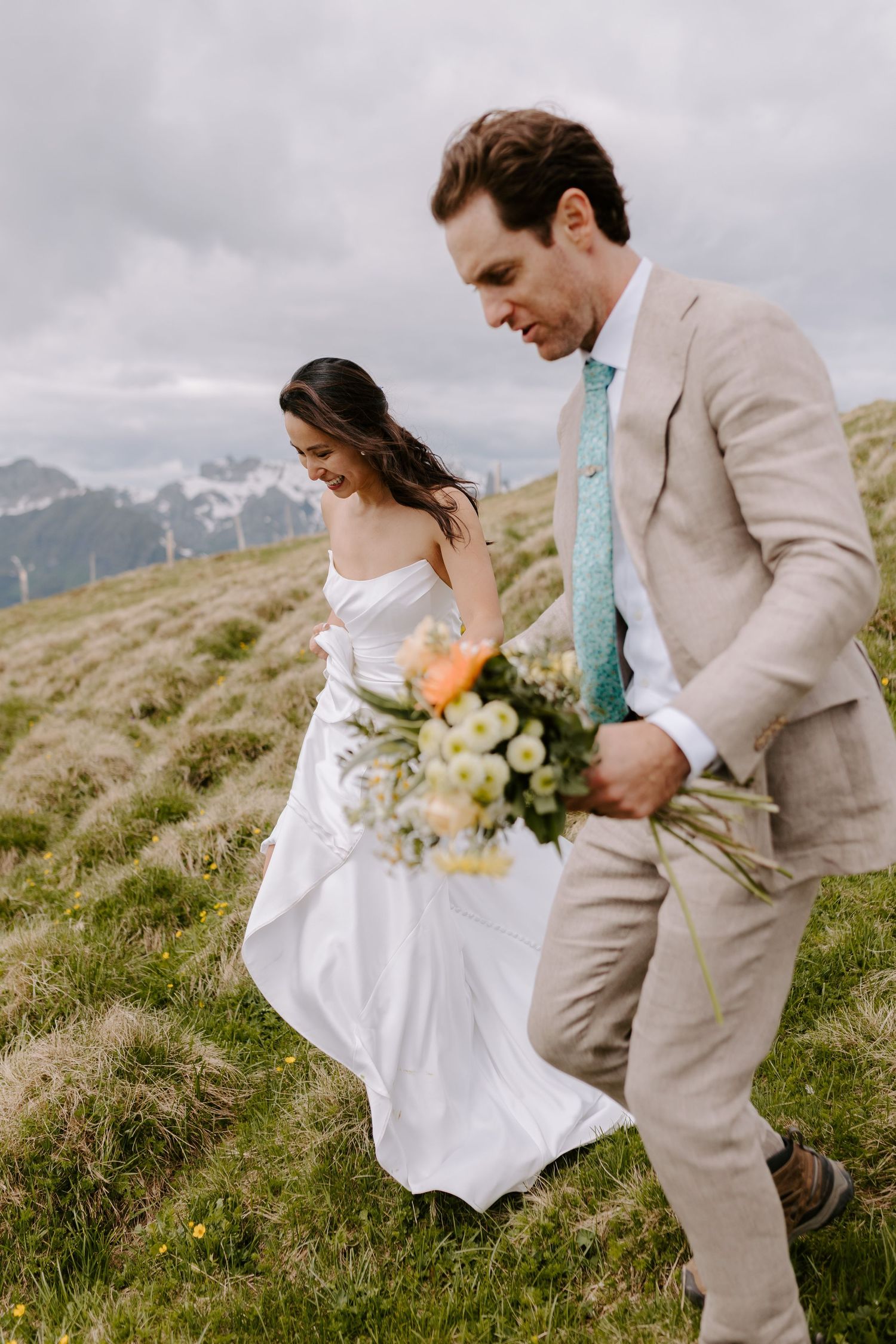 Newlyweds walking together on mountain hillside with bride holding floral bouquet.