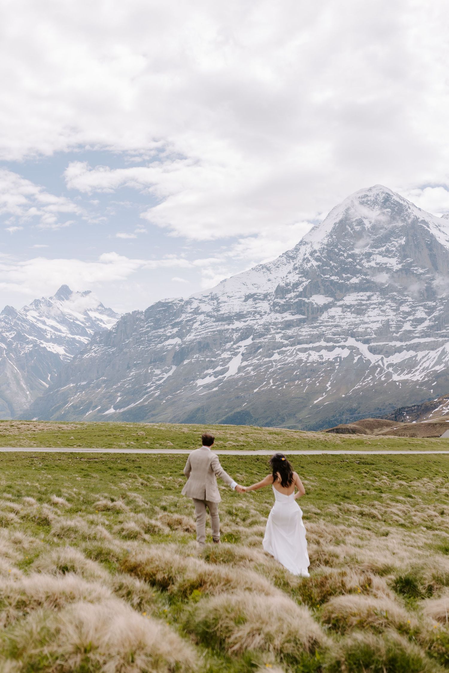 Couple holding hands in white wedding attire walking across meadow with snow-capped mountains in background.