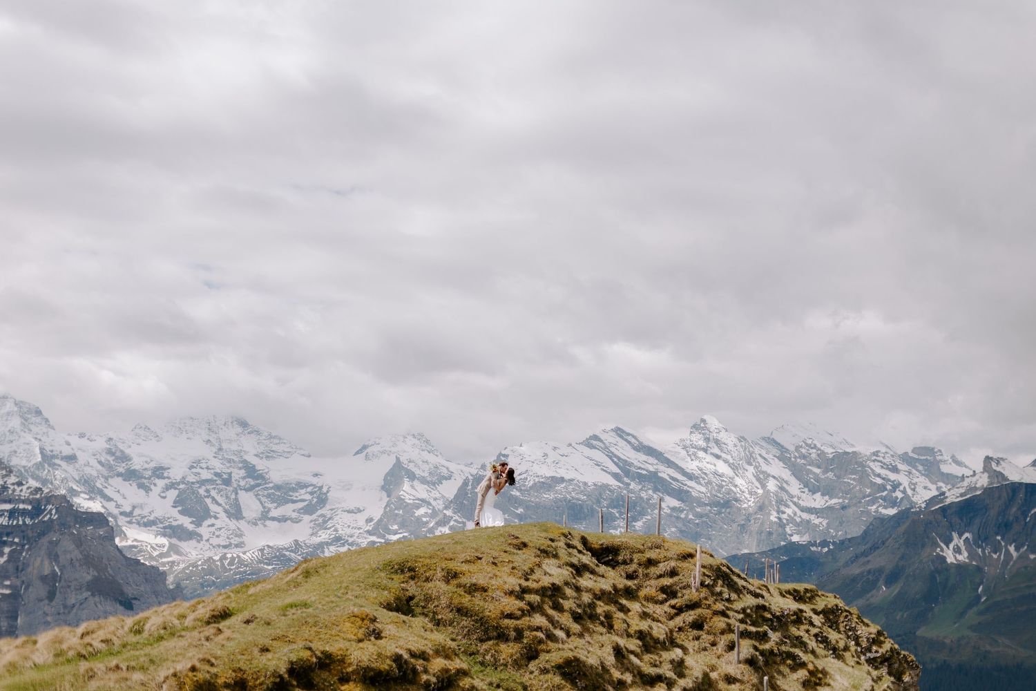 A scenic mountain panorama with snow-capped peaks and green slopes stretches across a cloudy horizon.