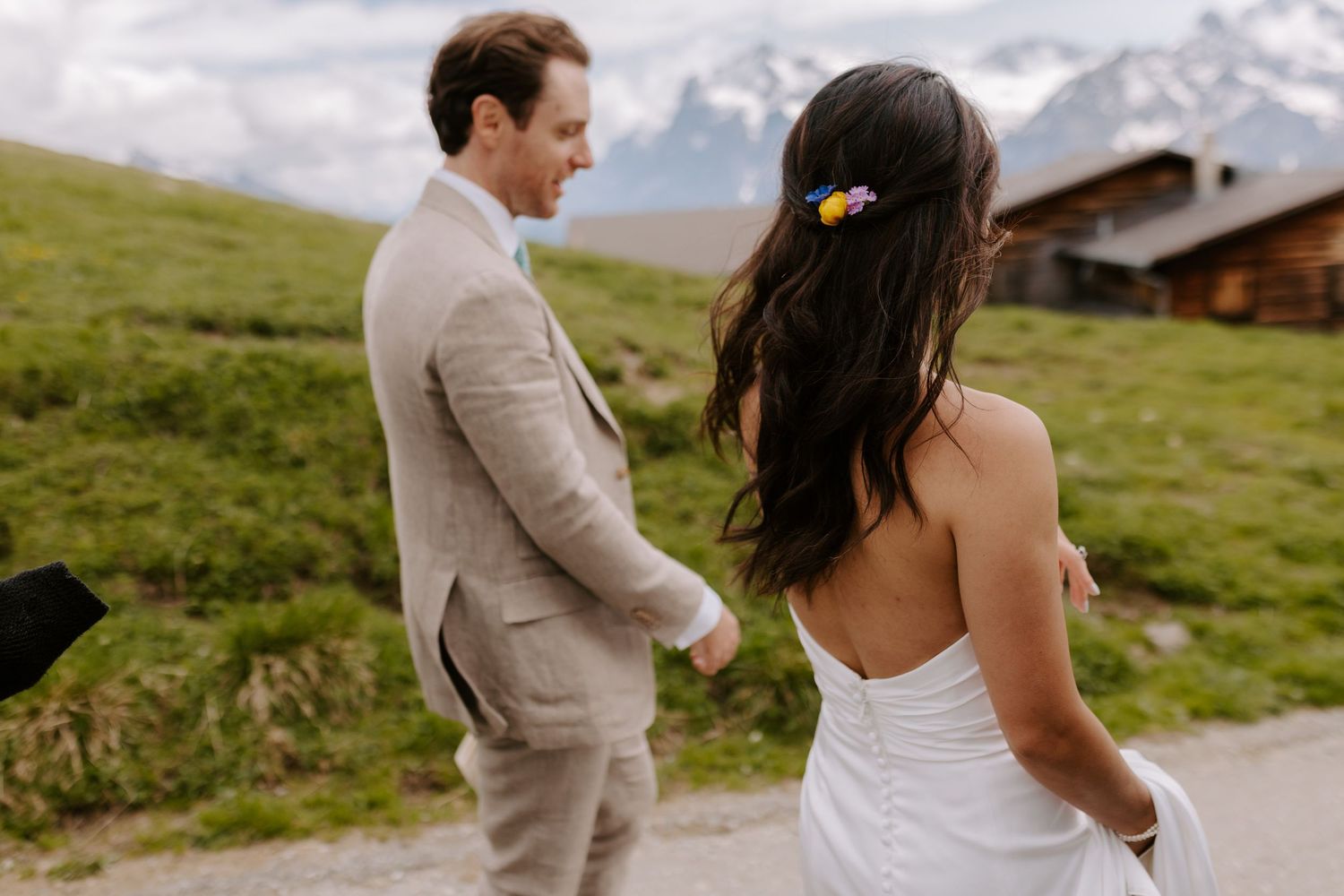 A couple in wedding attire walk together through a scenic alpine meadow with rustic mountain chalets in the background.