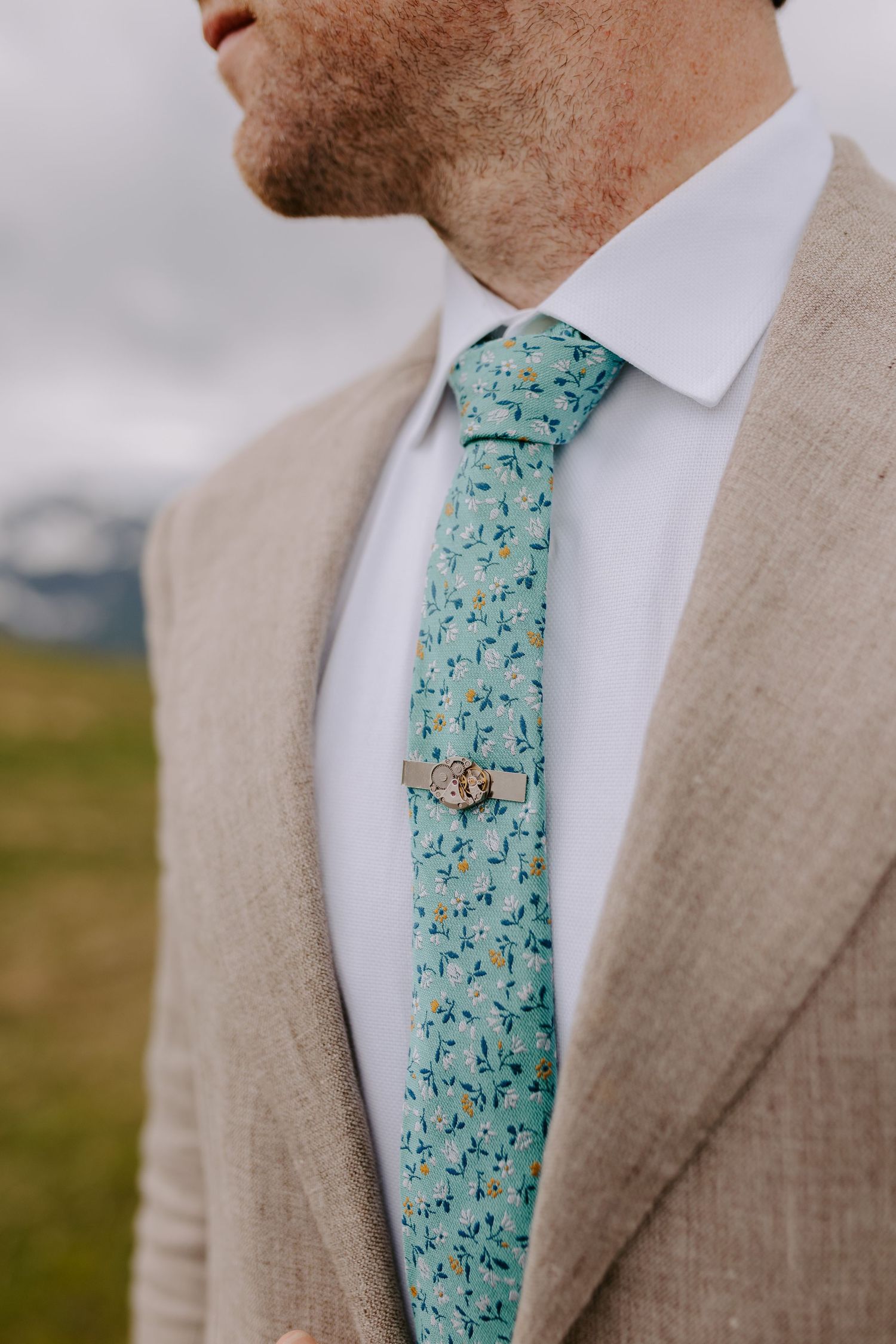 Close up of groom wearing light beige suit with mint green floral patterned tie and white dress shirt.