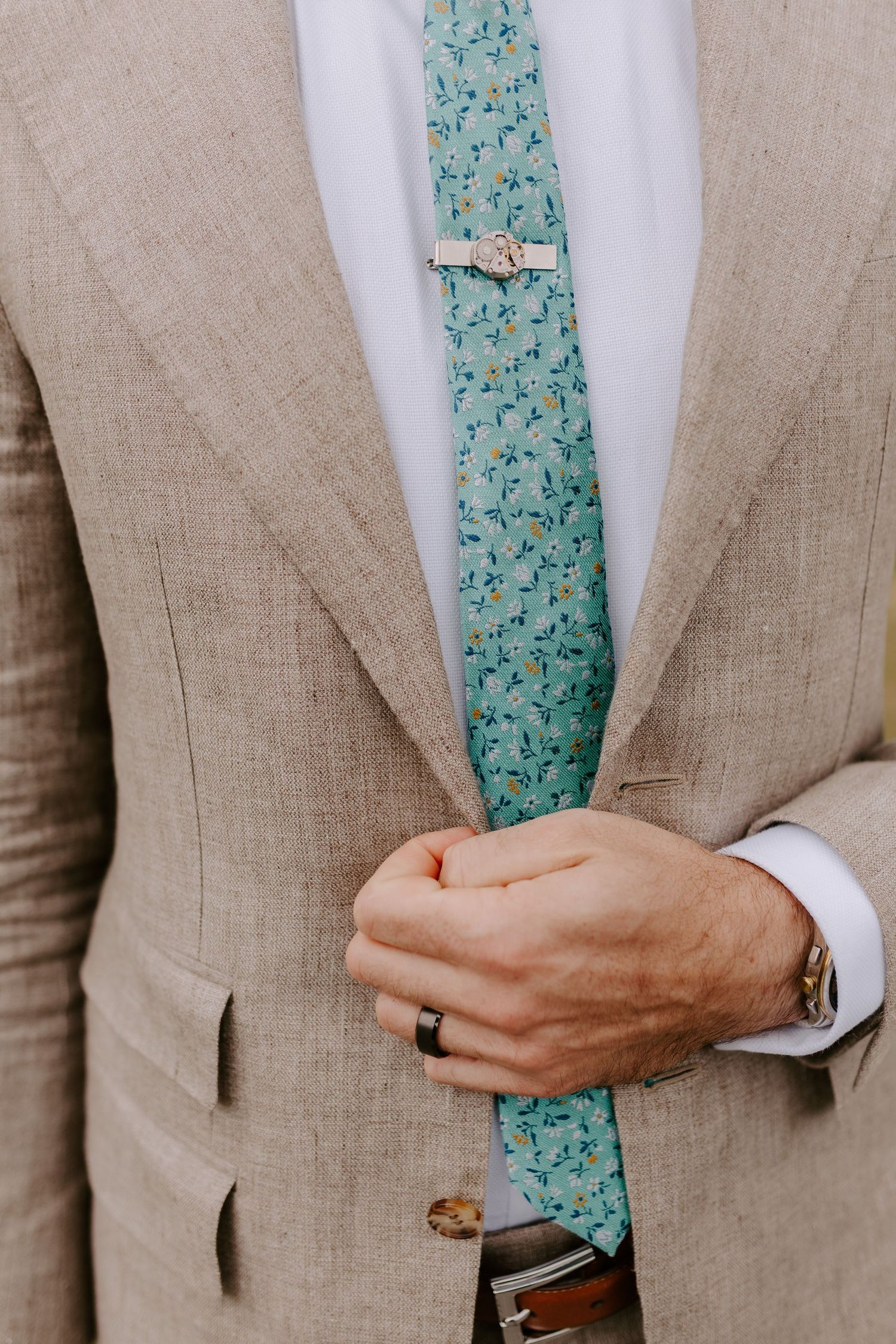 A person wearing a tan linen suit with a light blue floral tie adjusts their jacket sleeves and ring.