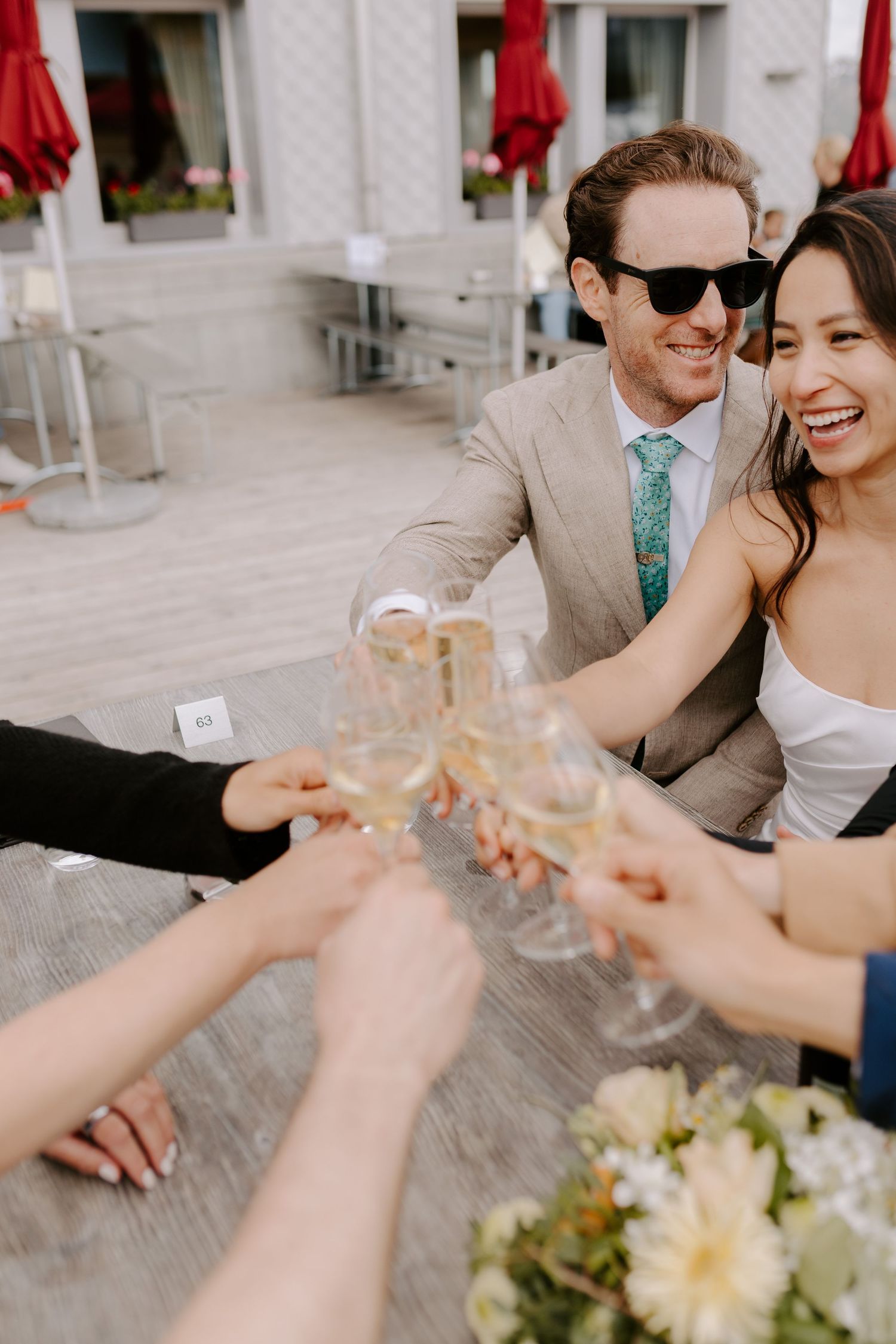 A couple toasts with guests at an outdoor reception with red umbrellas in the background.