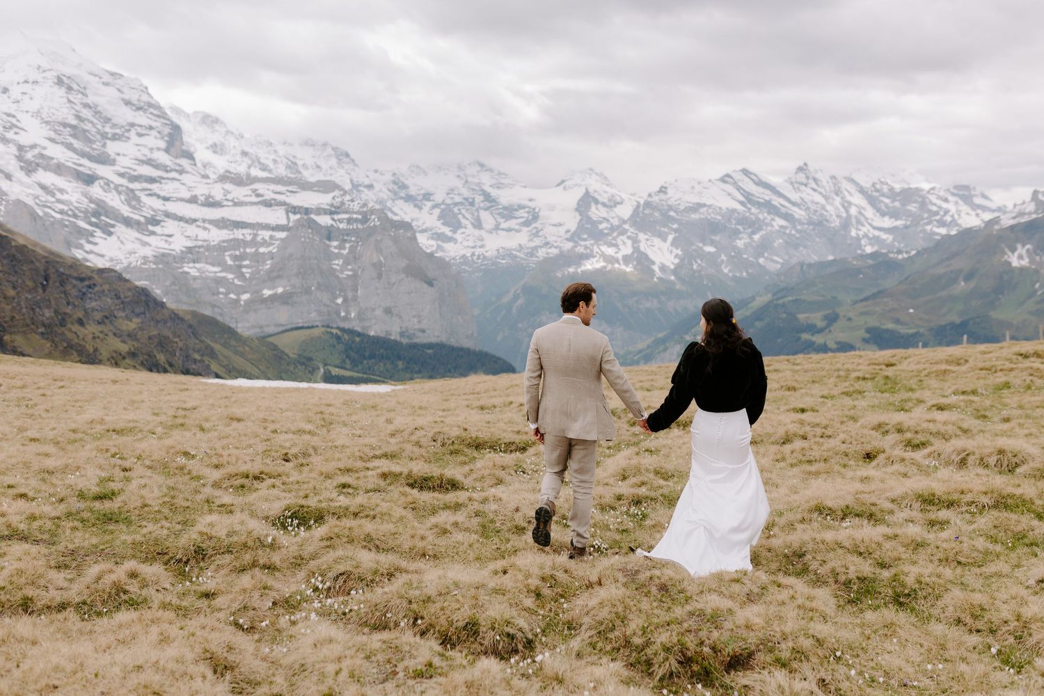Two people run hand in hand through a mountain meadow with snow-capped alpine peaks stretching across the horizon.