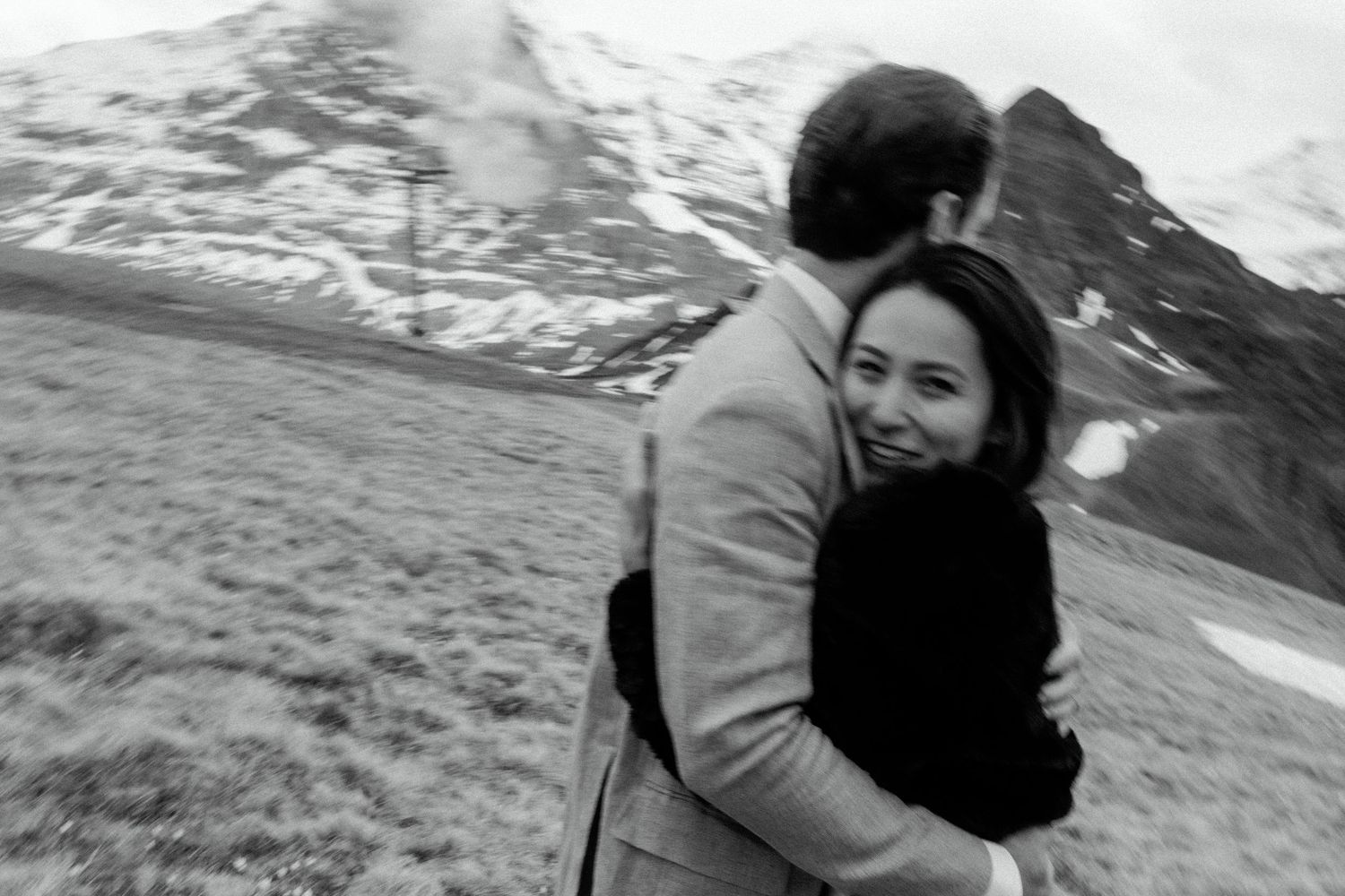Couple embracing on mountainside with snow-capped peaks in black and white.