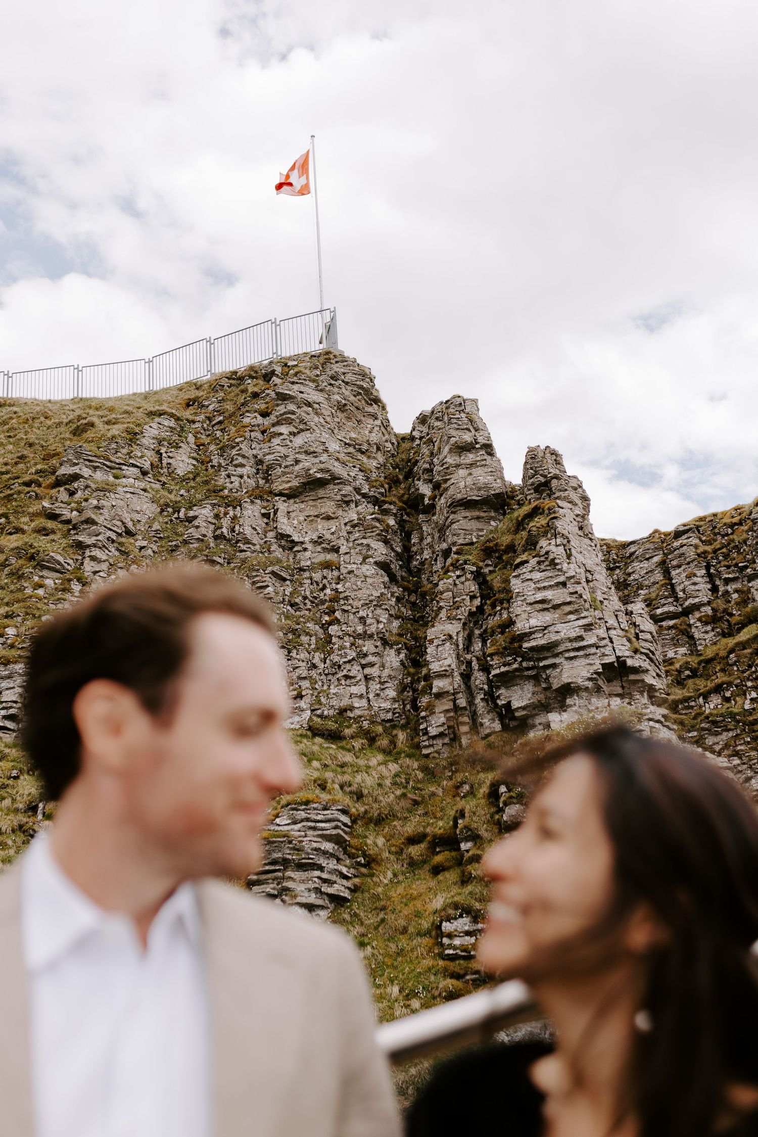 A Swiss flag waves atop rocky cliffs as a couple poses below during their mountain wedding.