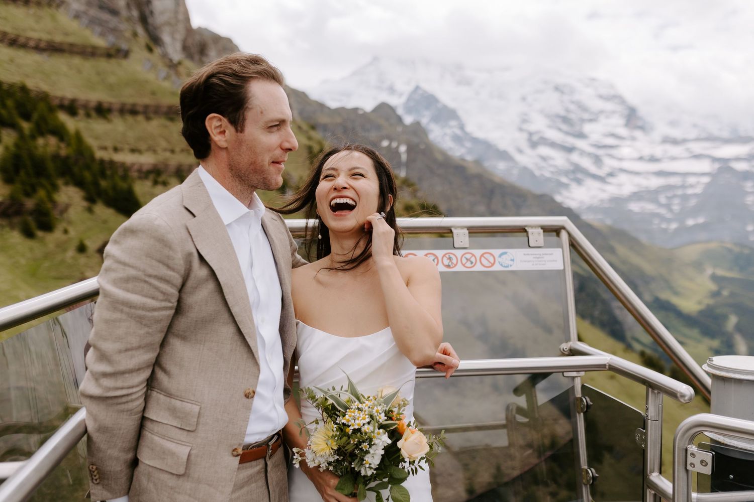 Newlyweds sharing an intimate moment on a viewing platform with majestic mountain vistas behind them.