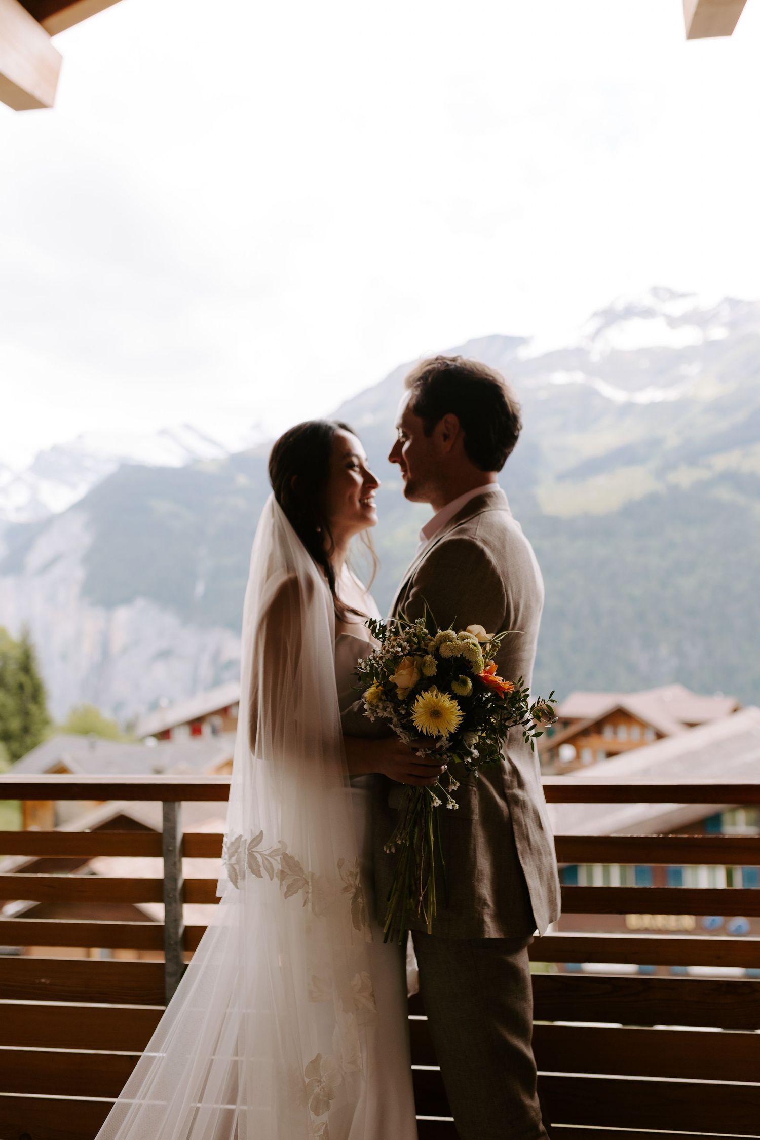 Wedding couple embracing on a wooden balcony with sunlit Alpine mountains in the backdrop.