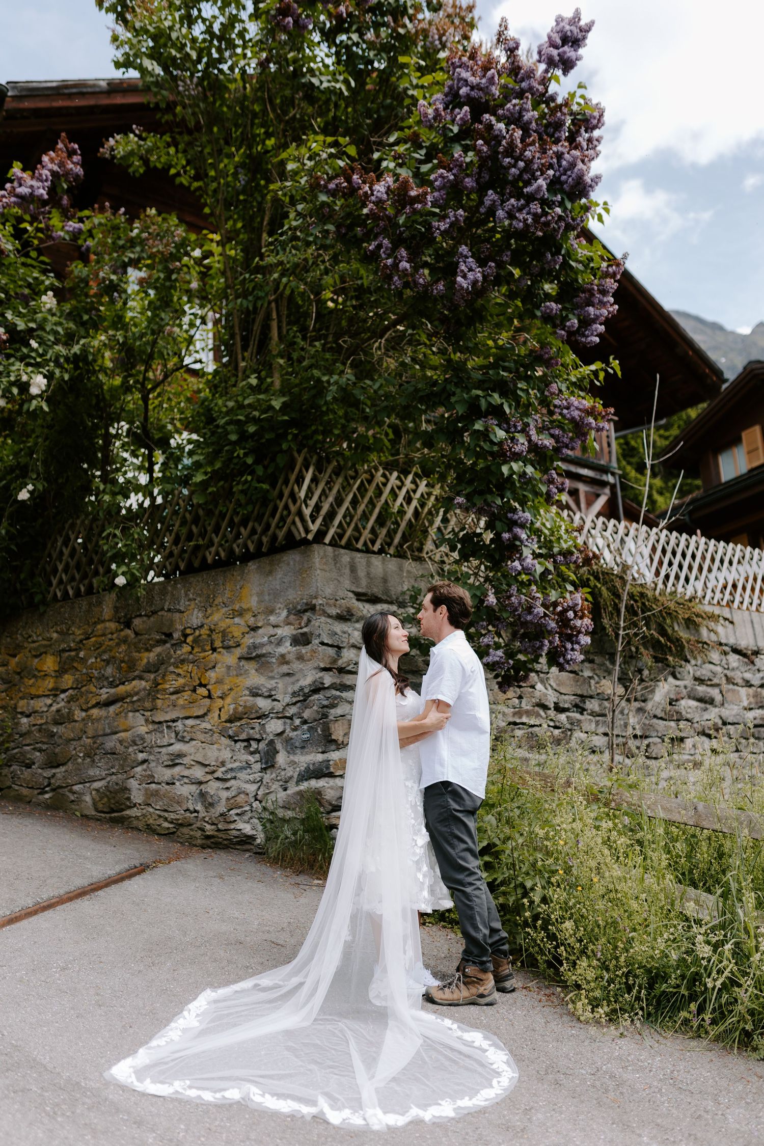 A couple in wedding attire embrace by a stone wall with blooming lilac flowers in a garden setting.