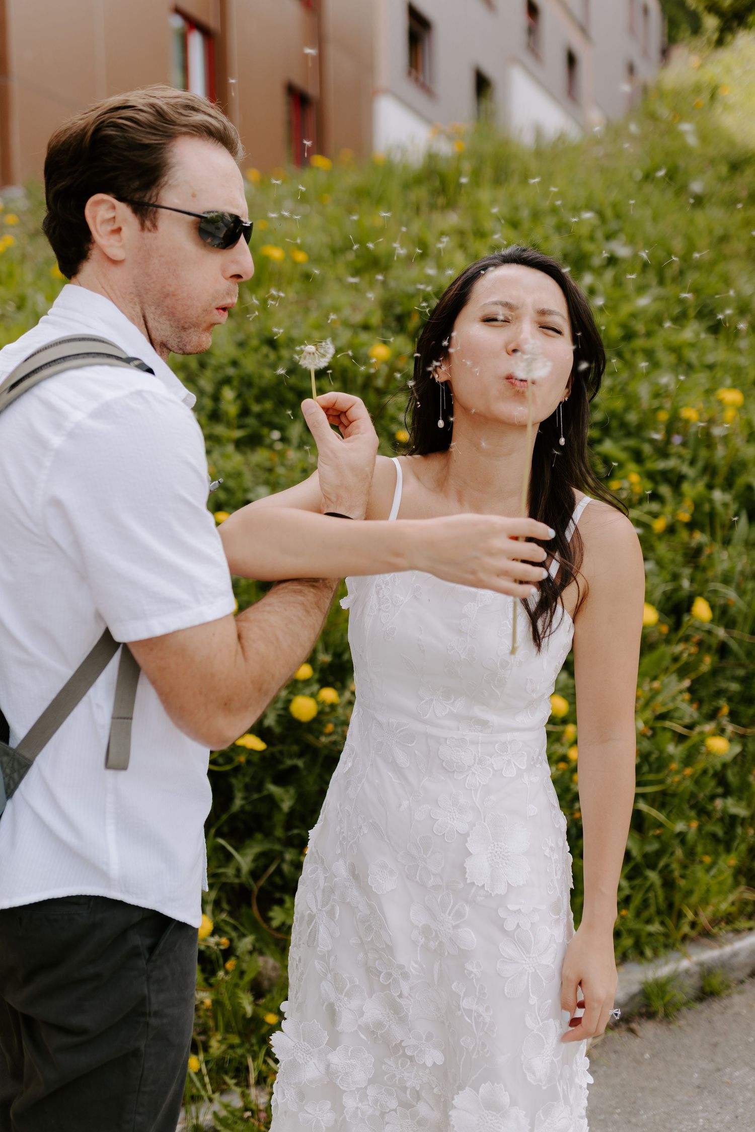 A couple playfully blows on dandelions in a flower-filled meadow during their wedding day.
