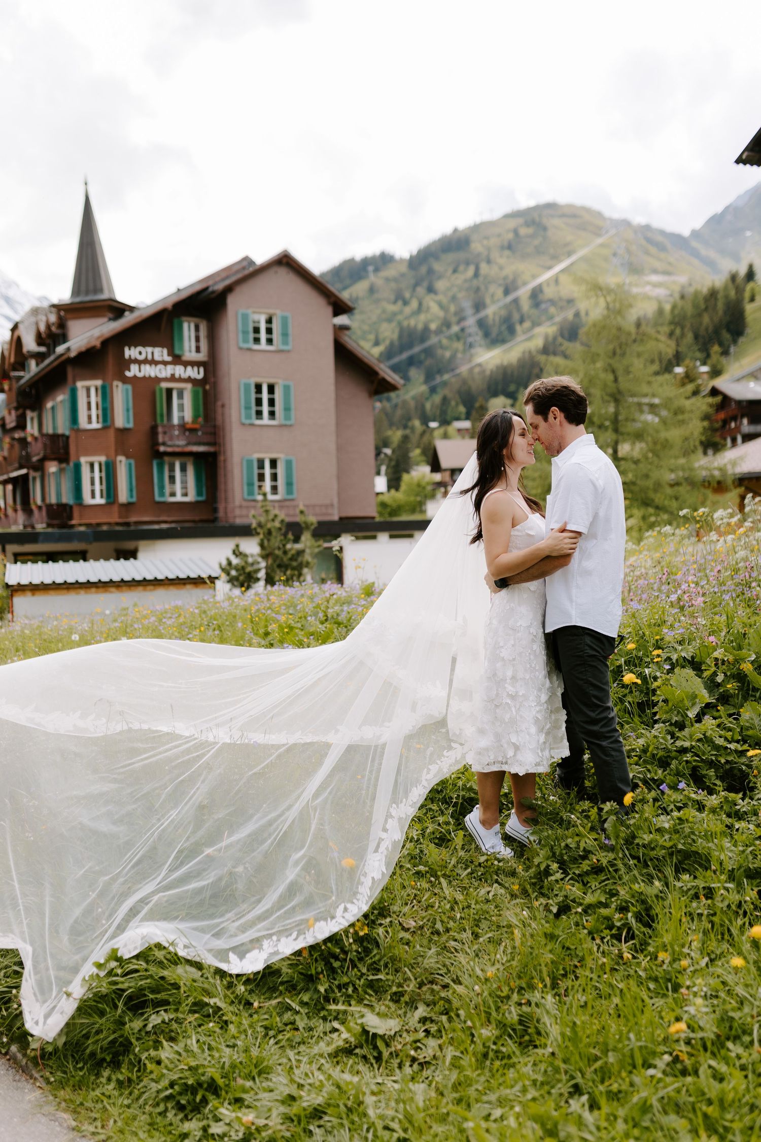 Wedding couple posing in front of alpine chalet with flowing white veil in mountain scenery.