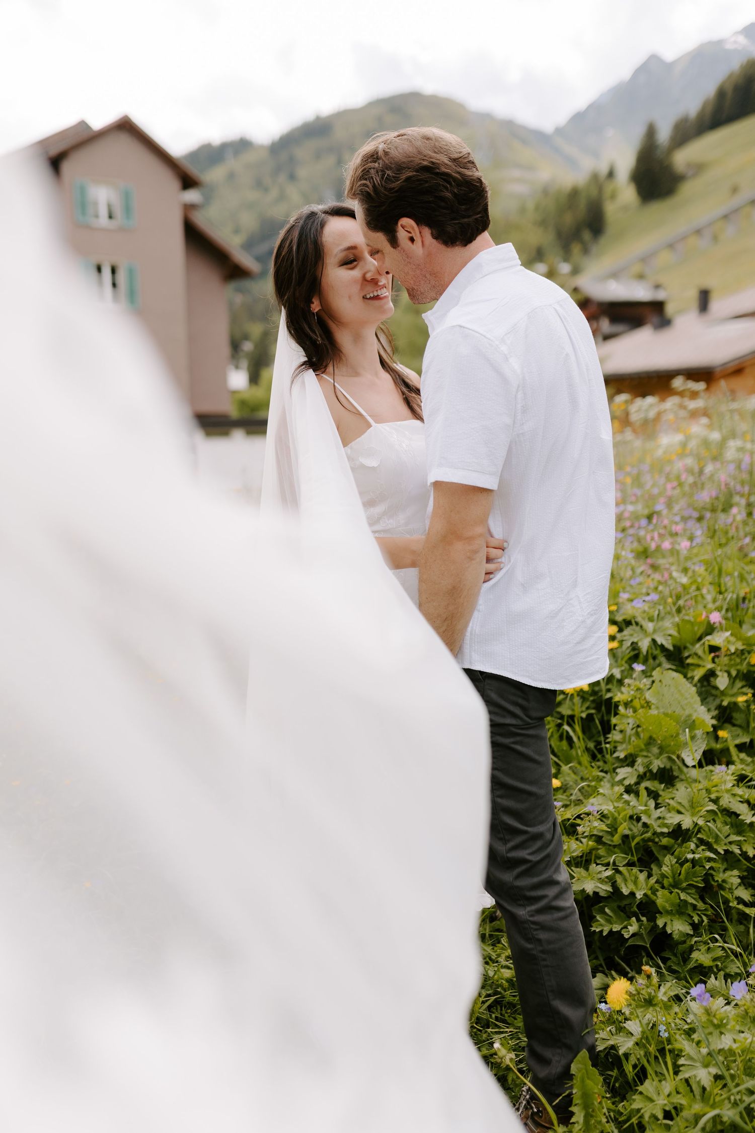 Intimate moment between couple in white against mountain backdrop with soft focus foreground.