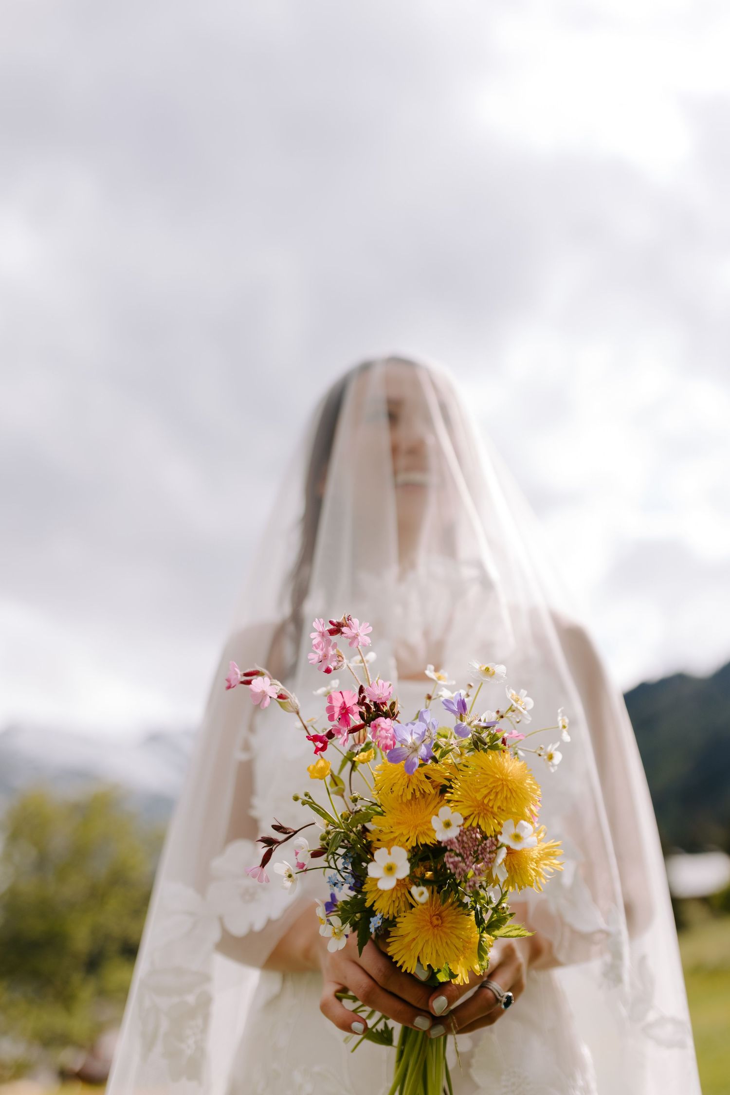 Bride in veil holds colorful wildflower bouquet with yellow daisies against cloudy mountain sky.