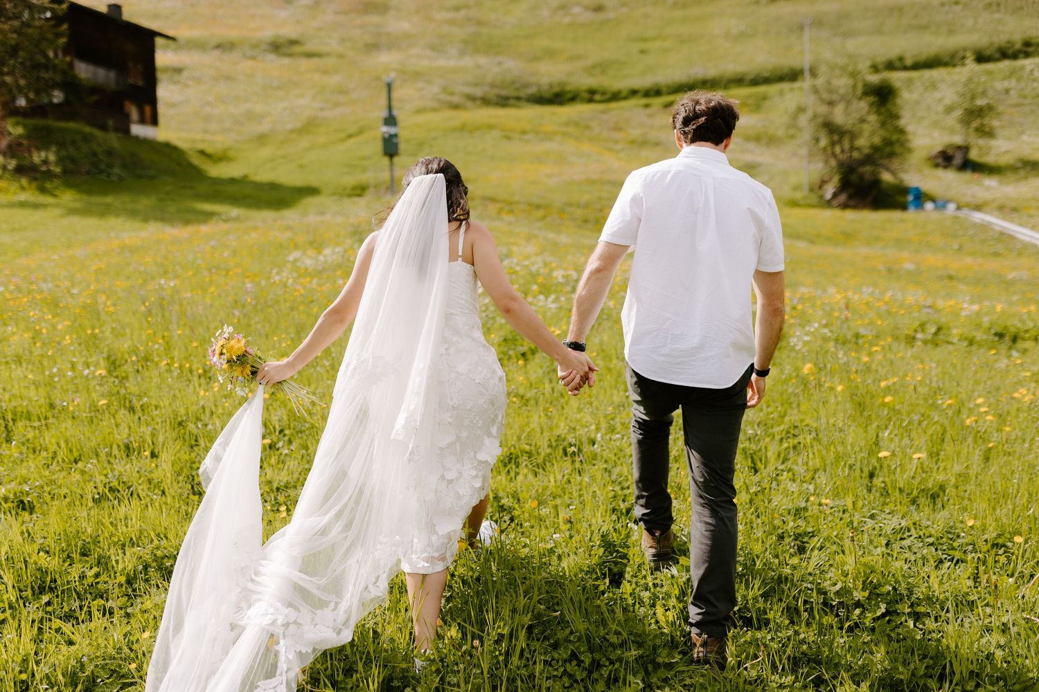 A couple walks hand in hand through a grassy meadow with the bride's veil flowing in the wind.
