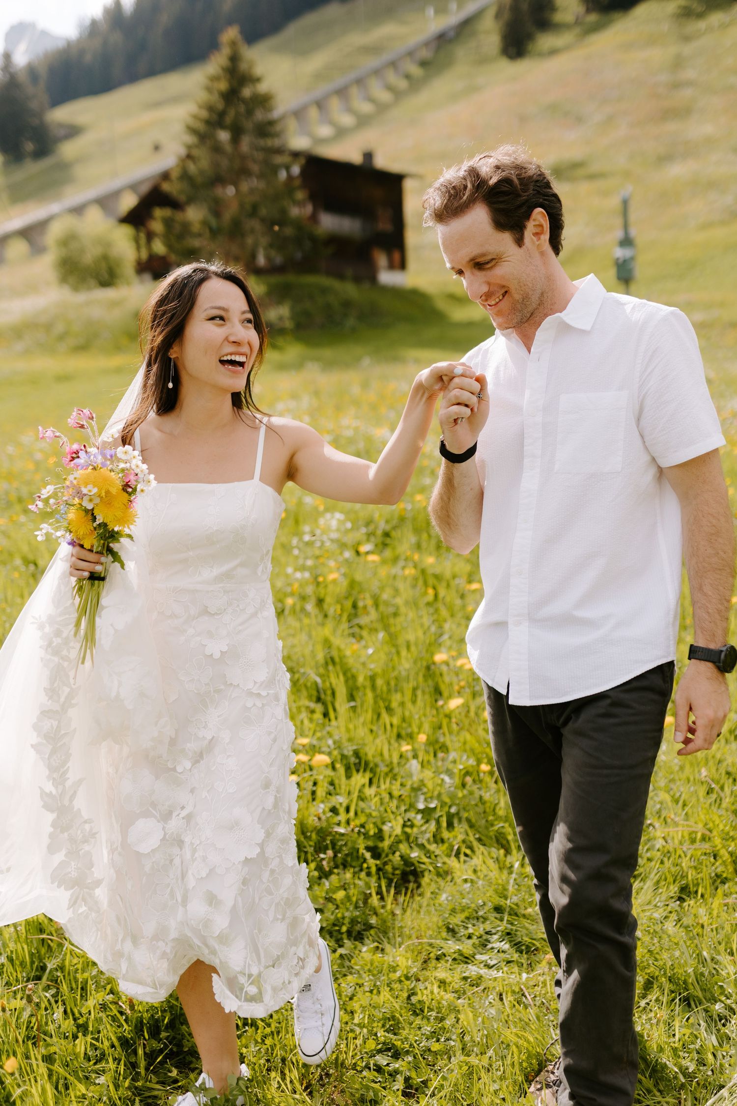 A joyful couple walks through a sunny alpine meadow with mountain chalets in the background.