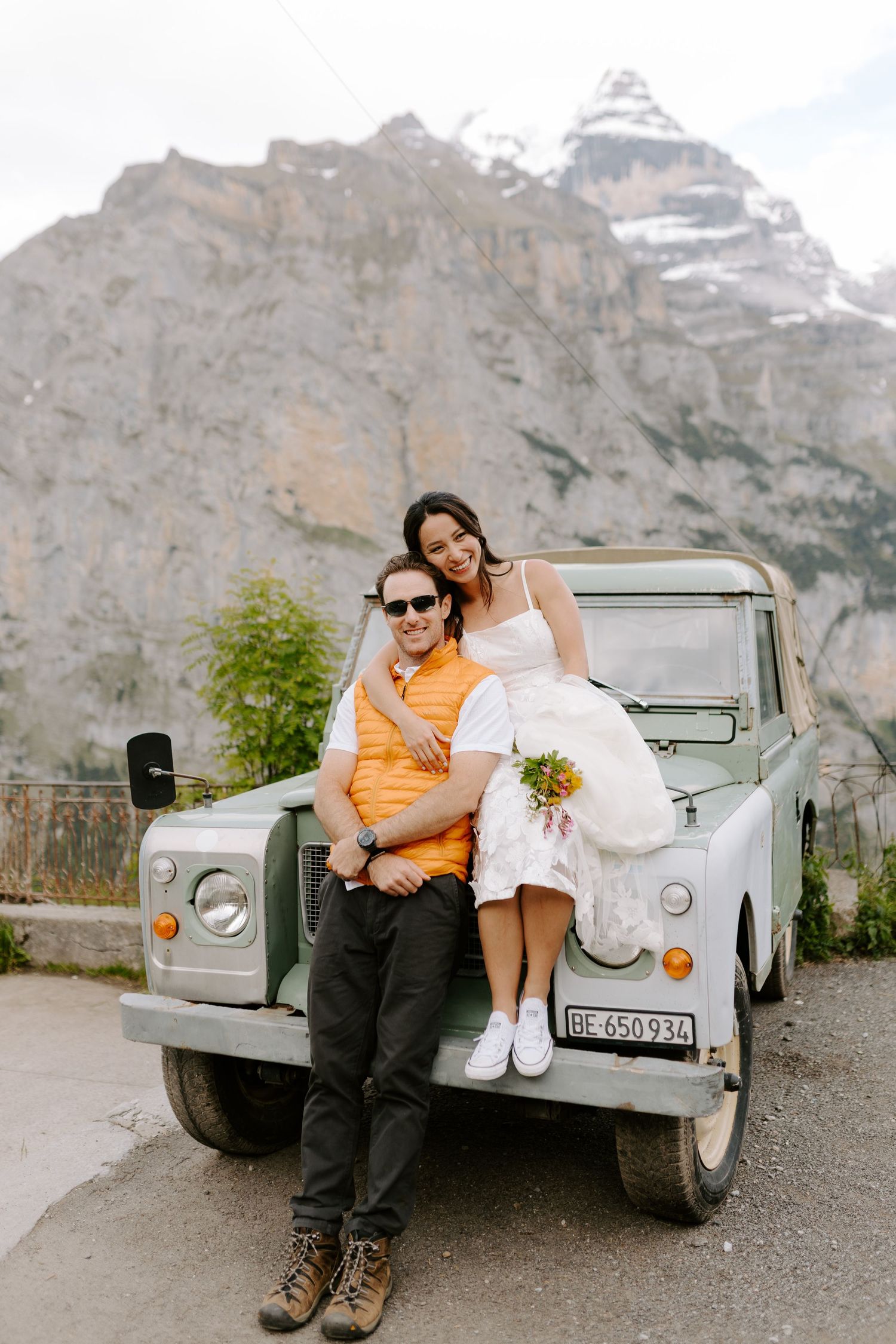 A couple poses with a vintage Land Rover against a dramatic mountain backdrop in the Alps.