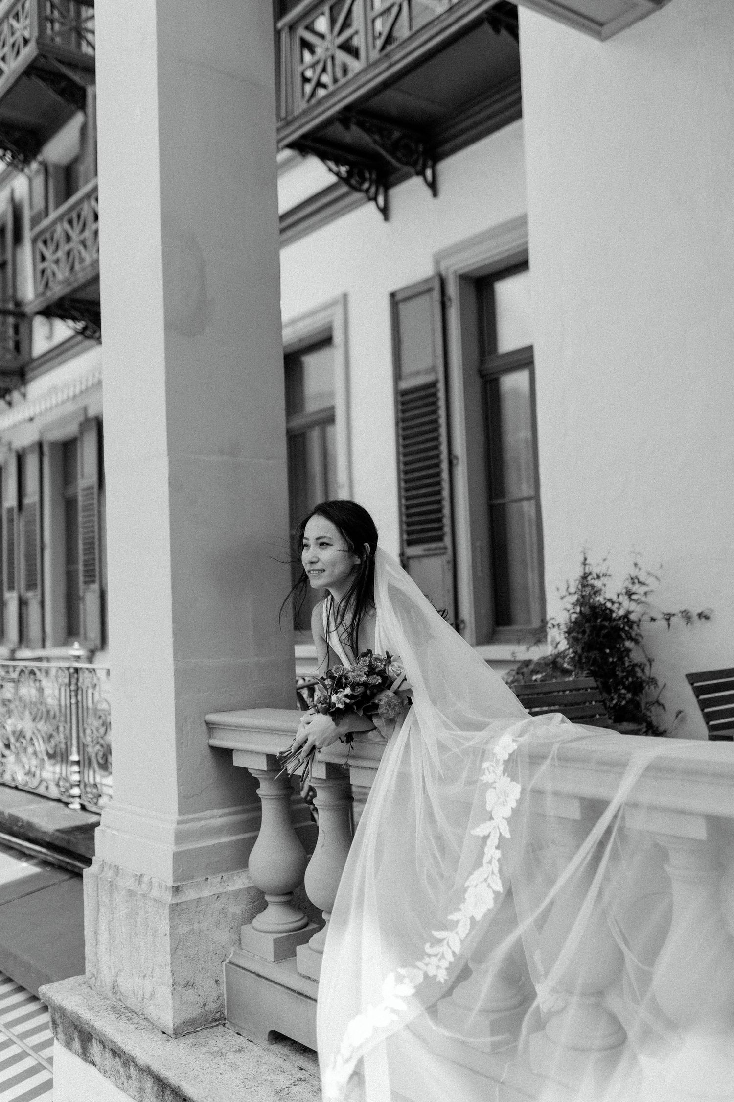 Black and white photograph of a joyful bride in a flowing veil on a classic building's front porch.