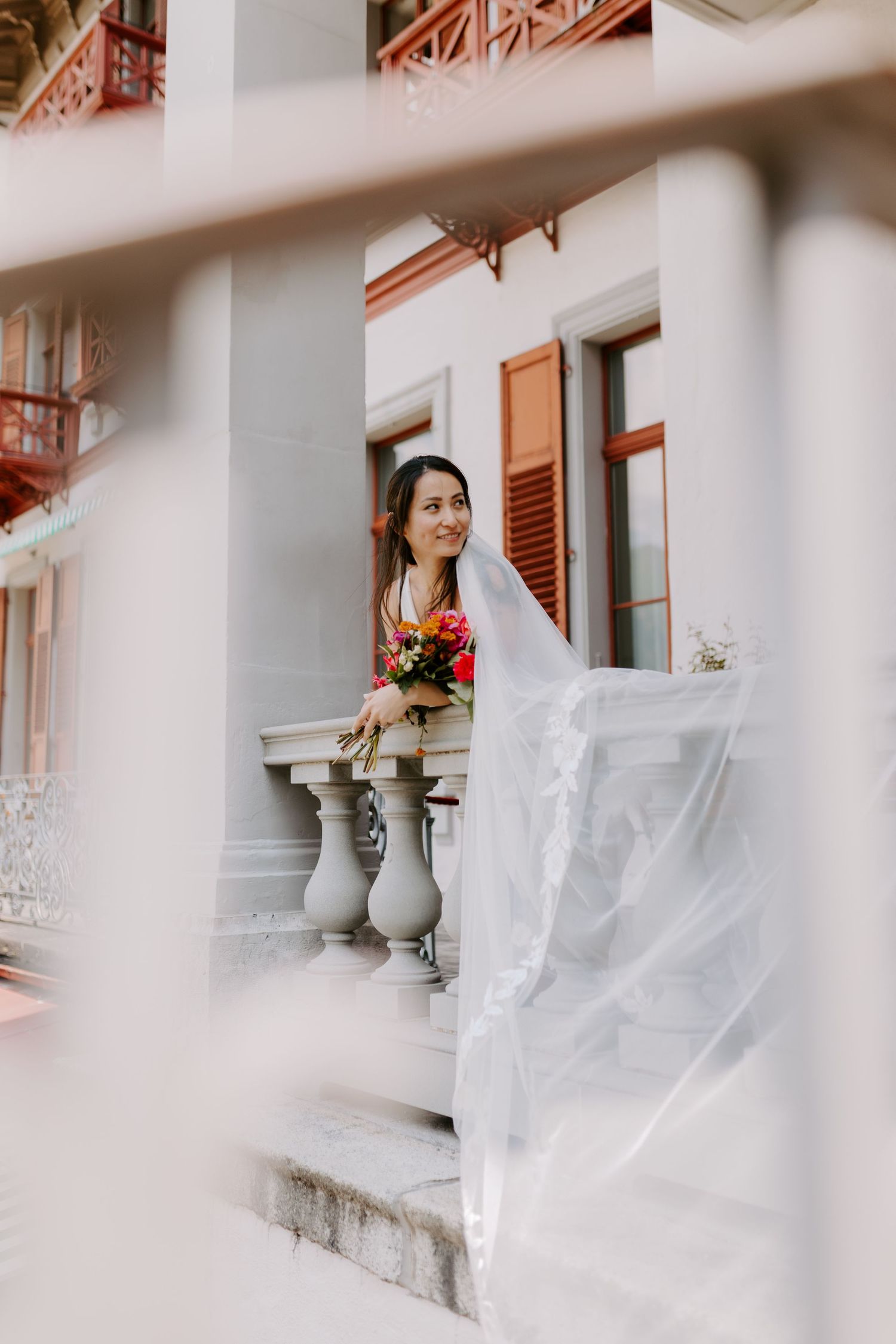 A bride in white dress holding flowers poses on classic balcony steps.