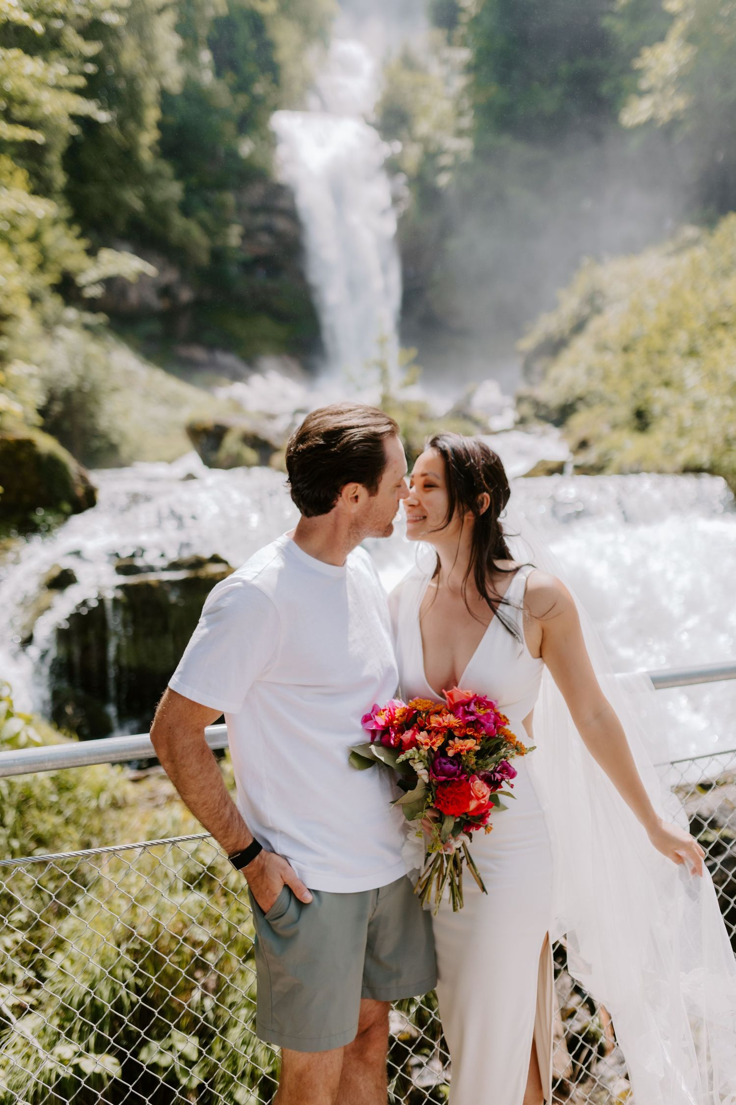 A couple poses together in front of a majestic waterfall holding bright pink flowers.