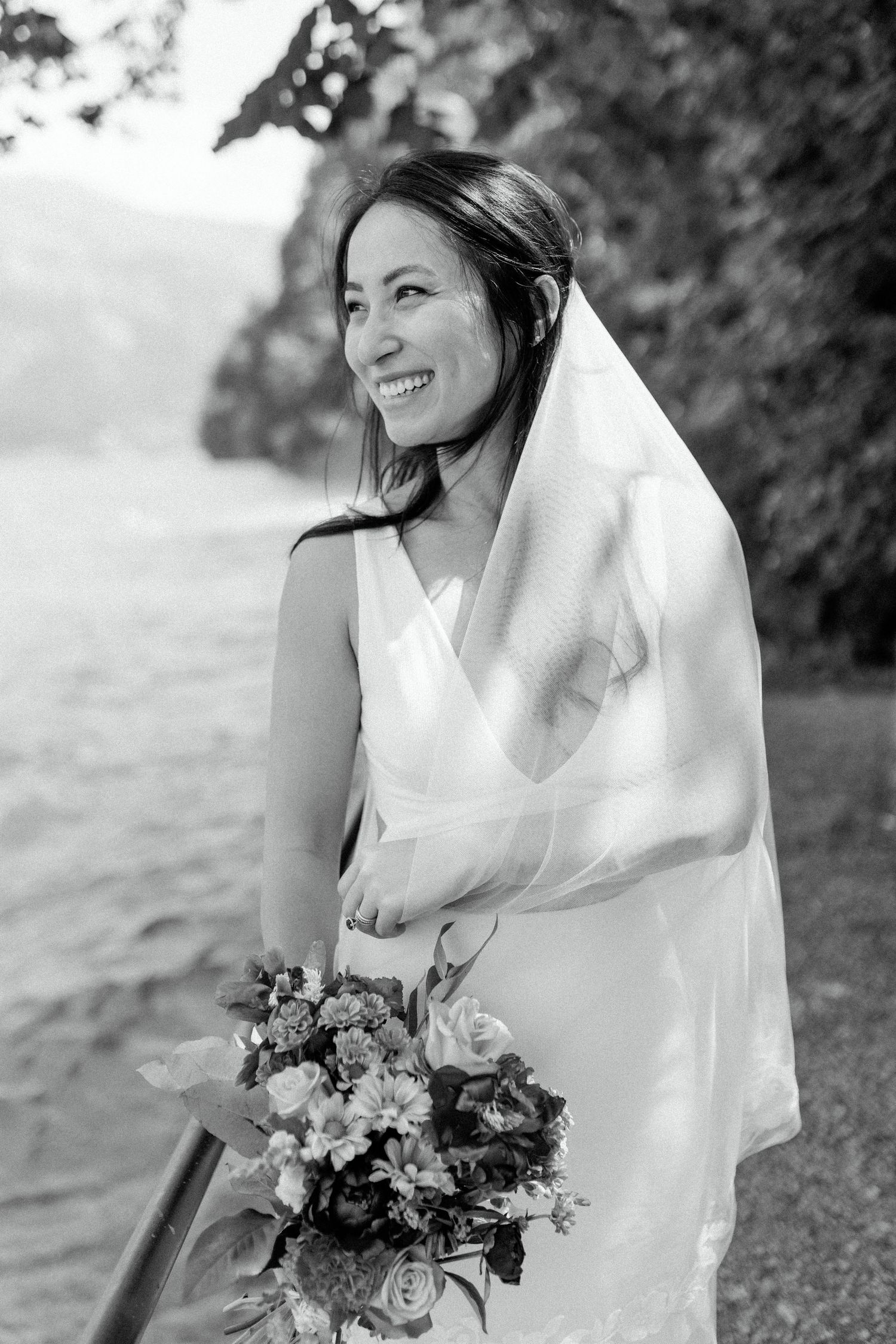 A bride in a white gown smiles joyfully while holding a floral bouquet against a lakeside backdrop.