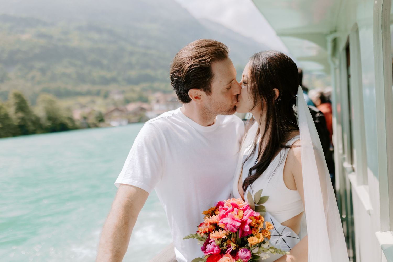 A couple shares a romantic kiss by a turquoise lake while holding a vibrant pink bouquet of flowers.