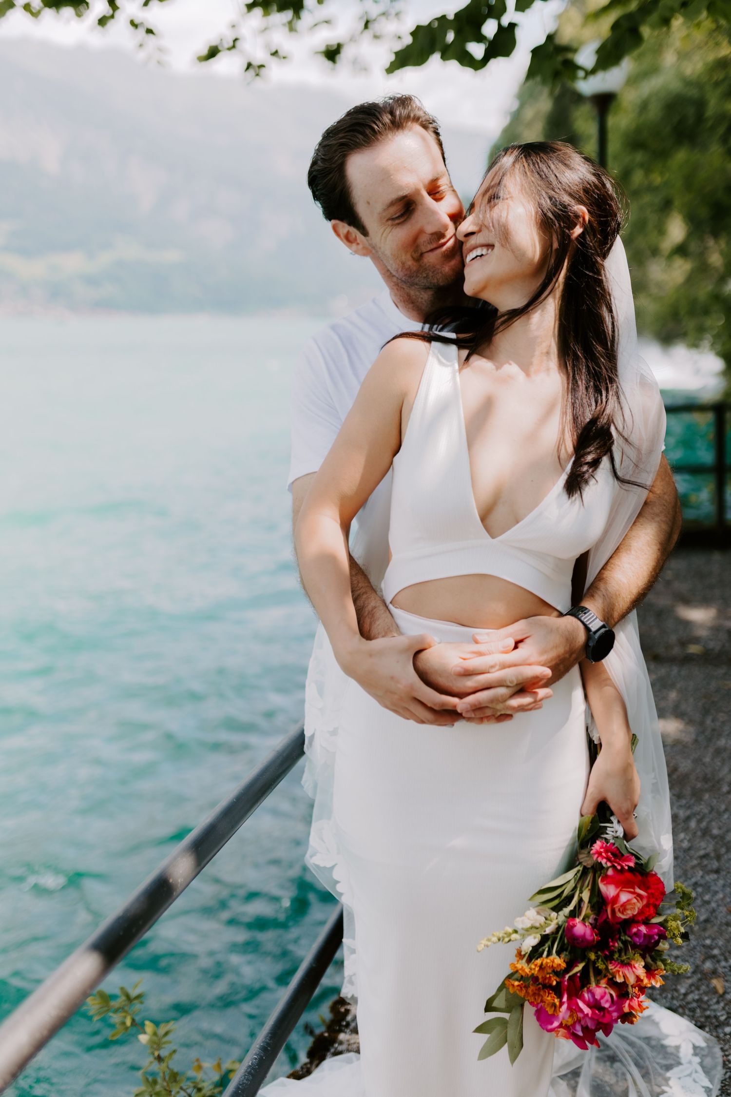 Romantic couple embracing by turquoise lake with bride holding vibrant red bouquet.
