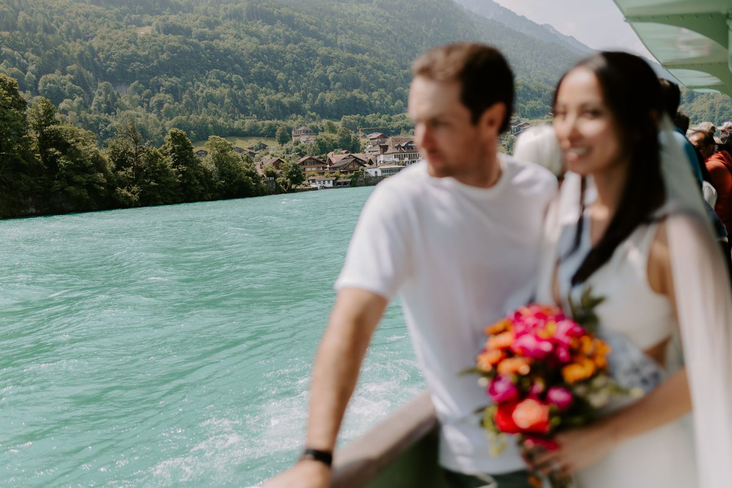 A couple poses near a turquoise lake with mountains and colorful flowers in view.