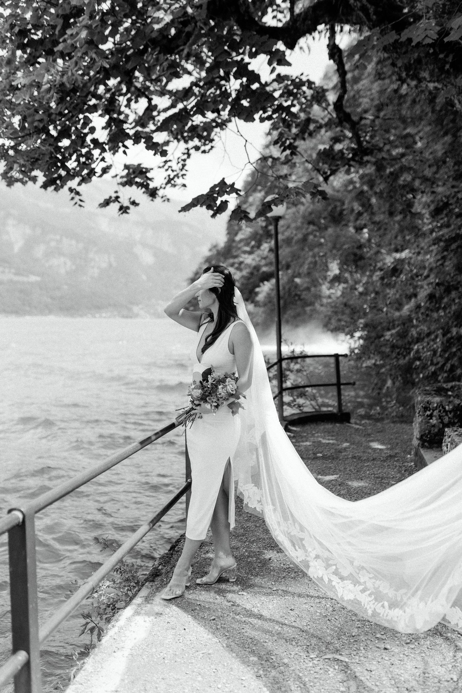 Dramatic black and white photo of bride with flowing veil by lakeside path.
