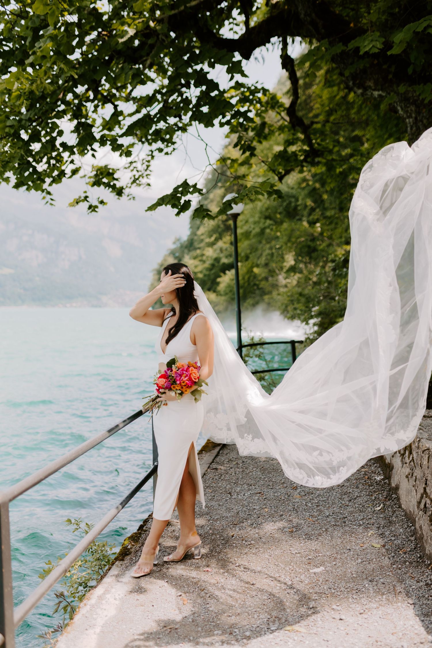 A bride in flowing veil poses dramatically on a lakeside path with mountains in the background.