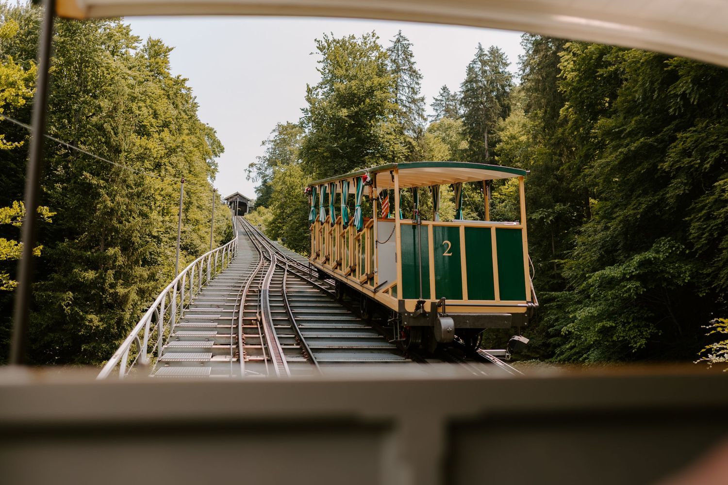 A vintage green funicular railway car climbs uphill through a forested mountain track.