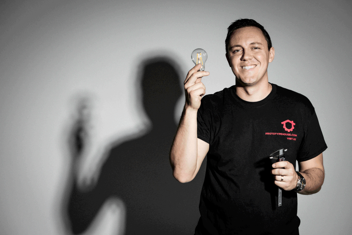 A person in a black t-shirt holds up a light bulb while casting a shadow against a gray background.