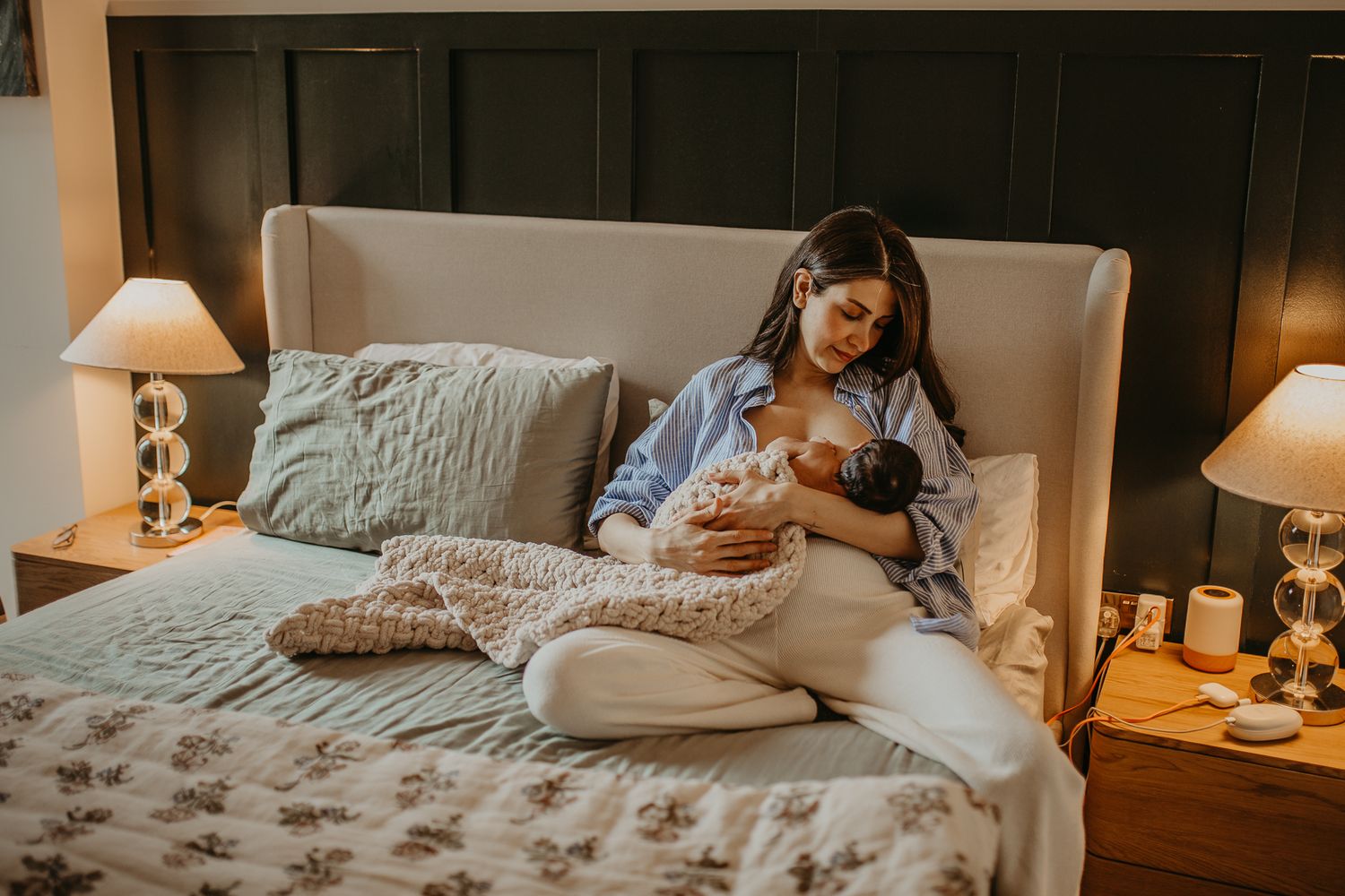 A nursing mother sits comfortably on a bed with white bedding and decorative pillows while lamplight creates a warm ambiance.